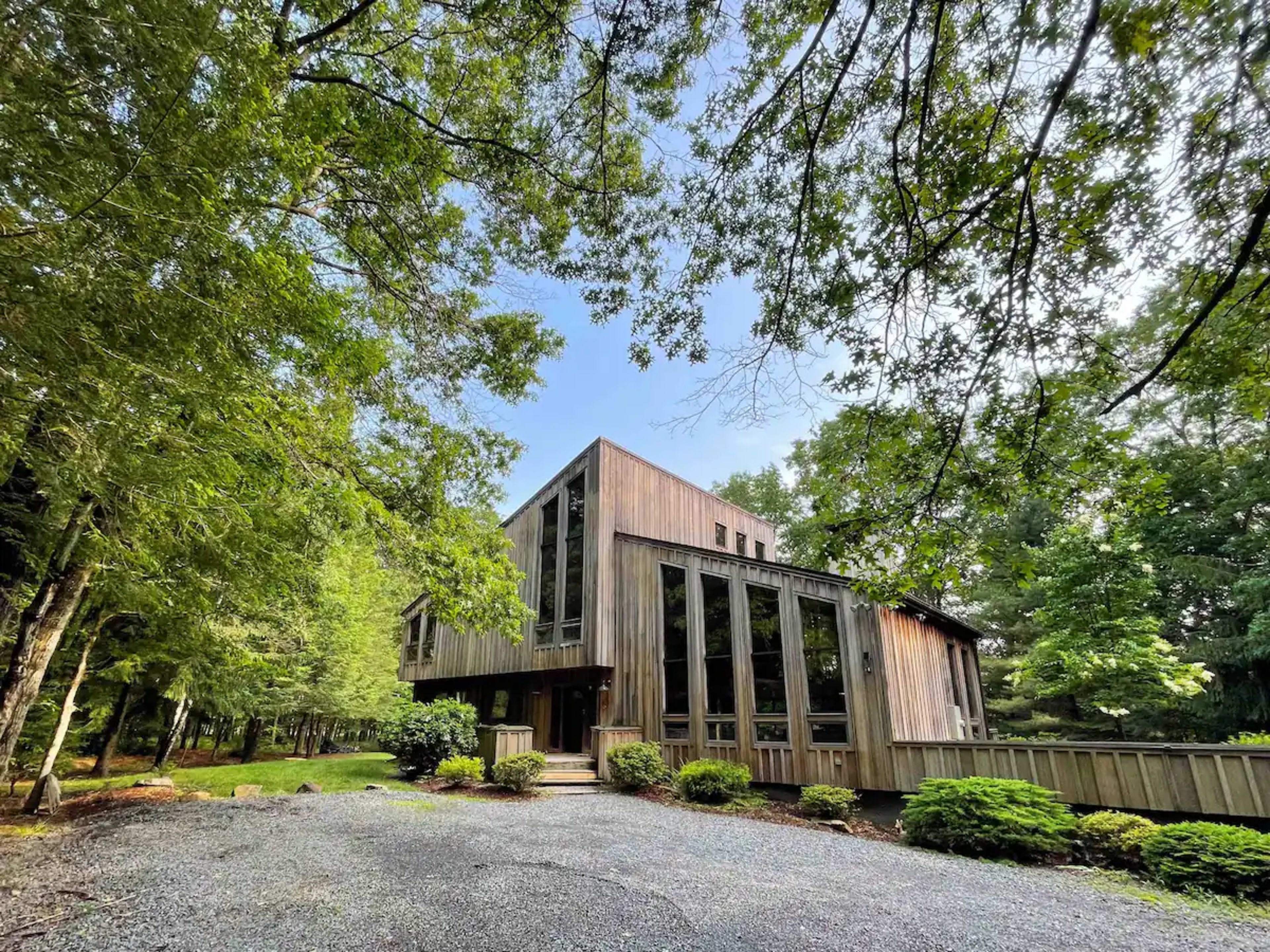 A wooden house with large glass windows is surrounded by trees and a gravel driveway.