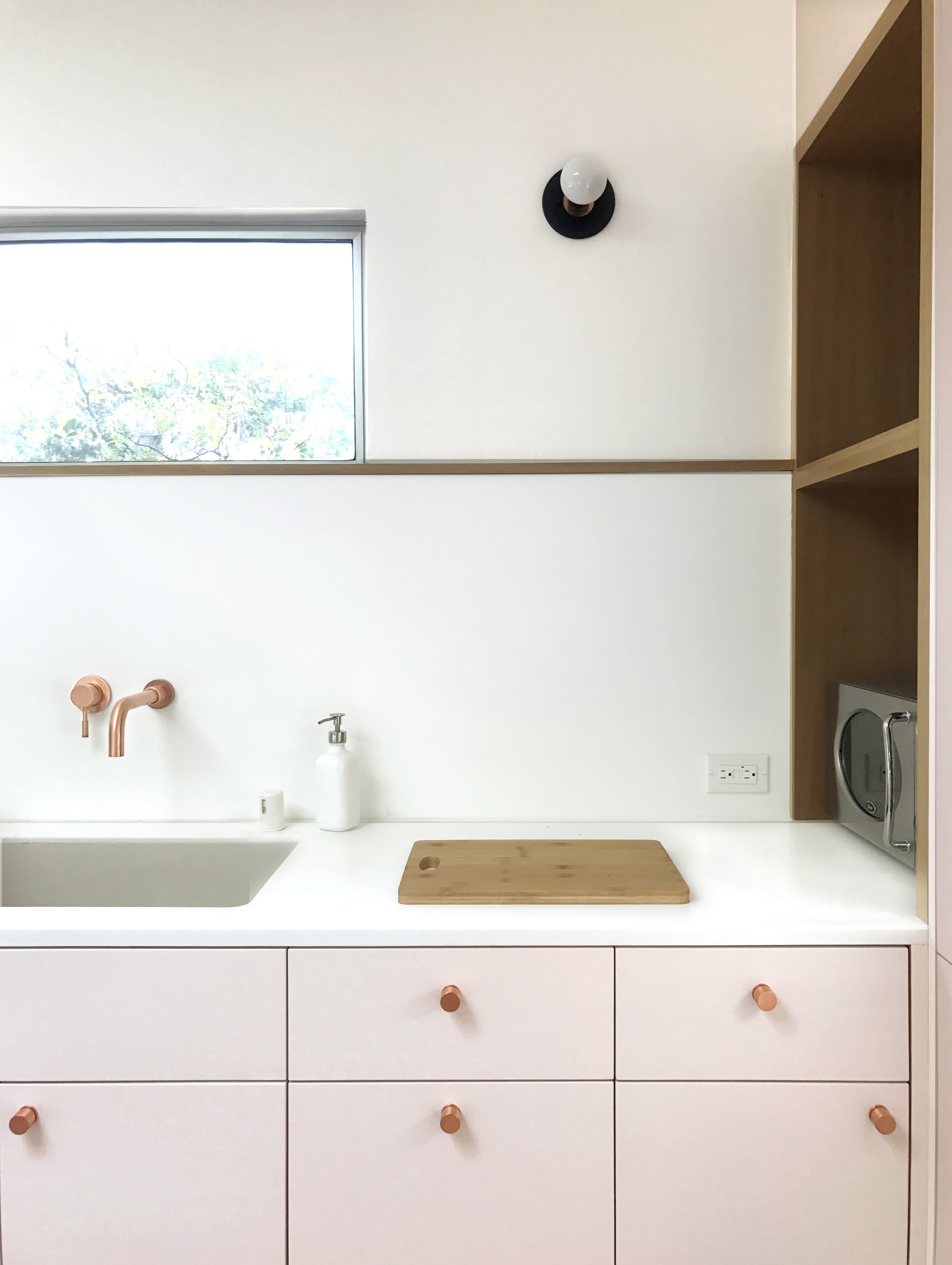 A modern kitchen features a white countertop with a wooden cutting board, a sink with rose gold faucets, and minimalist cabinetry.