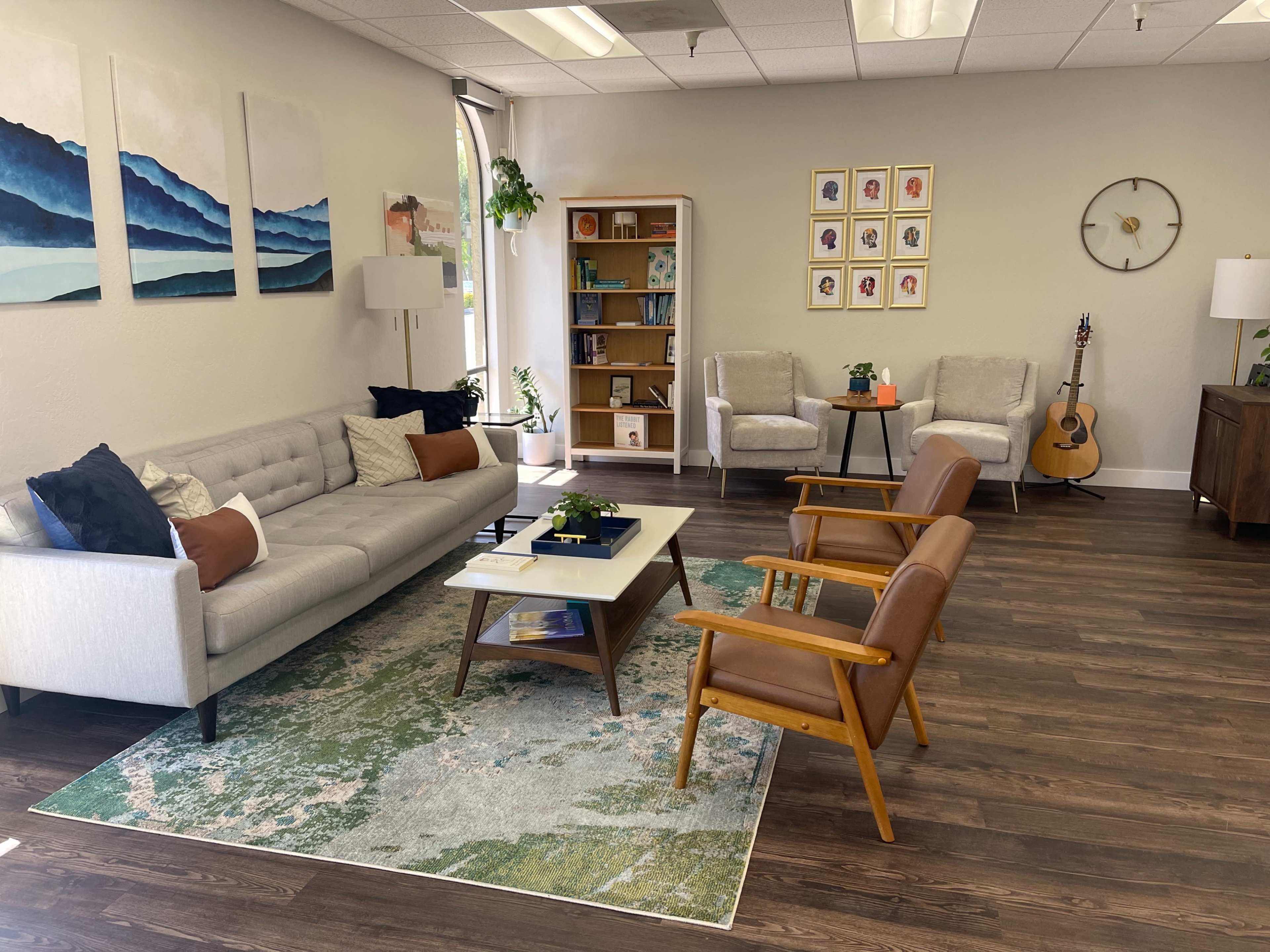A modern reception area with a light-colored sofa, wooden chairs, a coffee table, bookshelves, and decorative wall art.