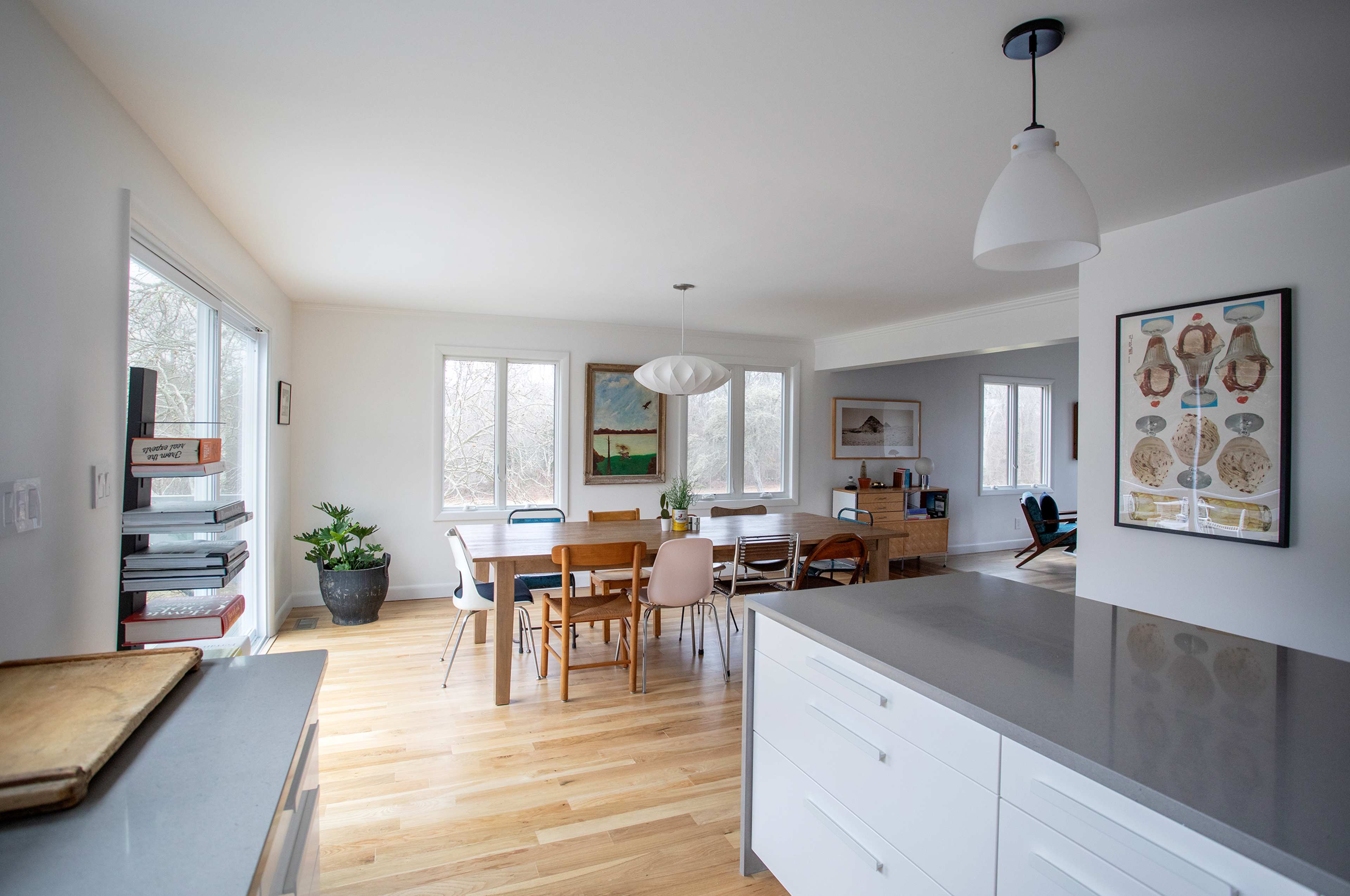 A modern kitchen opens to a dining area with wooden furniture and large windows overlooking a wooded view.