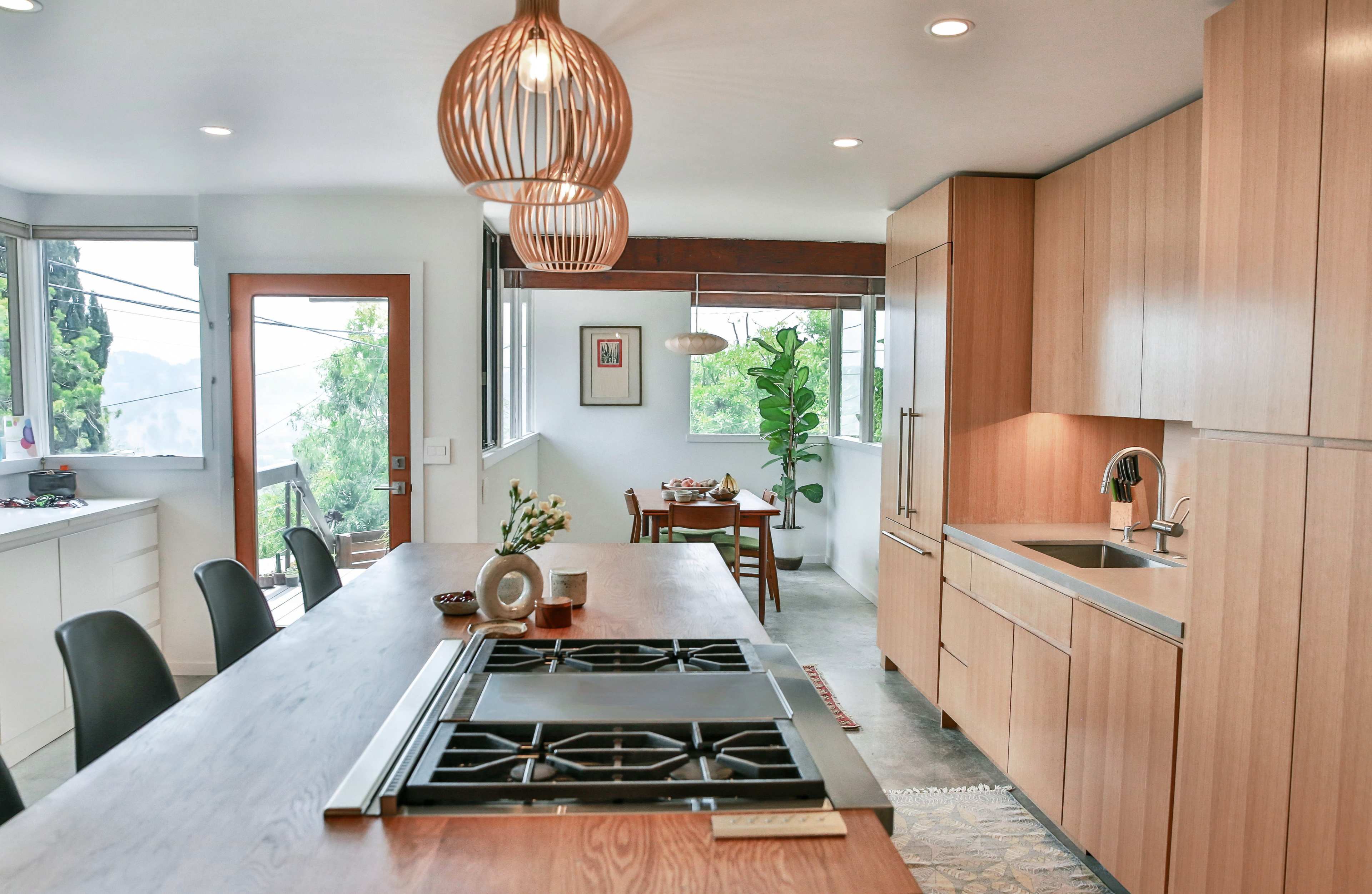 The image depicts a modern kitchen featuring wooden cabinetry, a central island with a stovetop, and a dining area adjacent to a window that overlooks greenery.