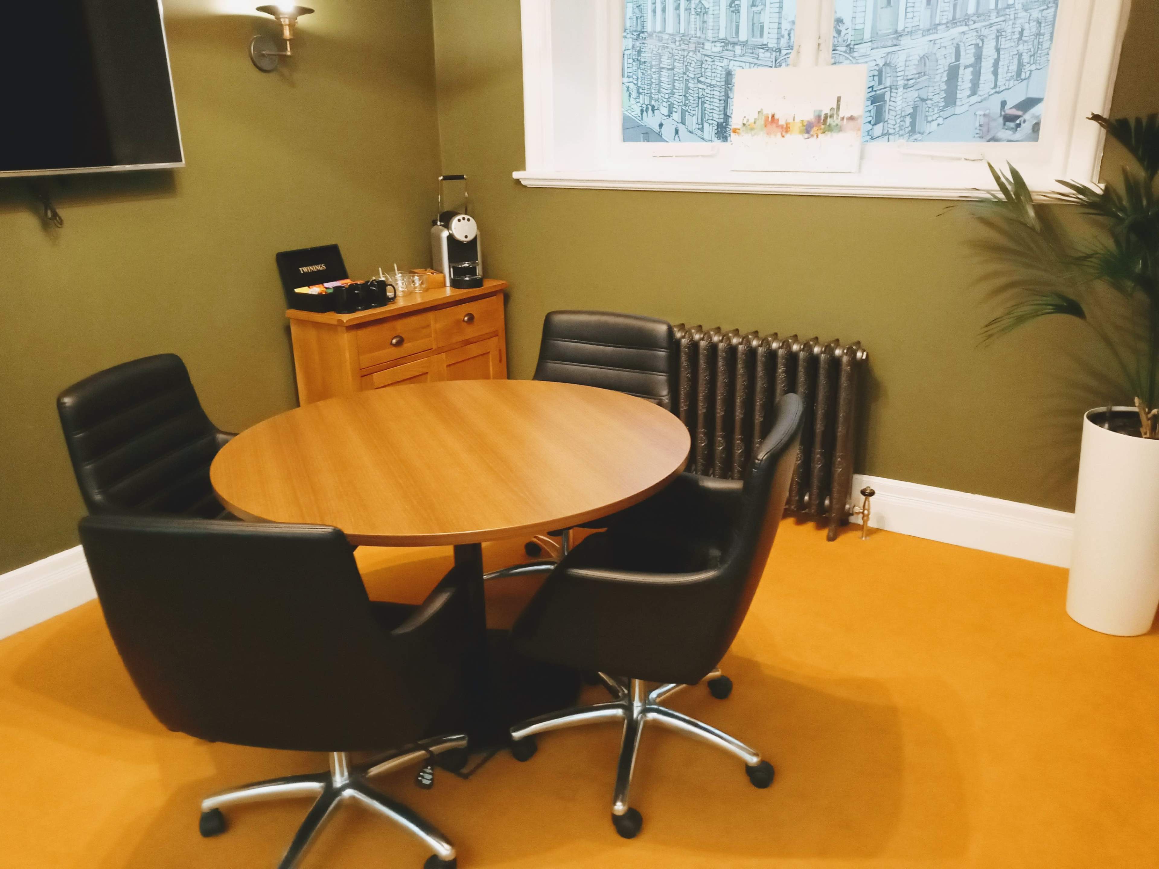 A small meeting area with a round wooden table surrounded by four black swivel chairs, a radiator, and a side cabinet against a green wall.