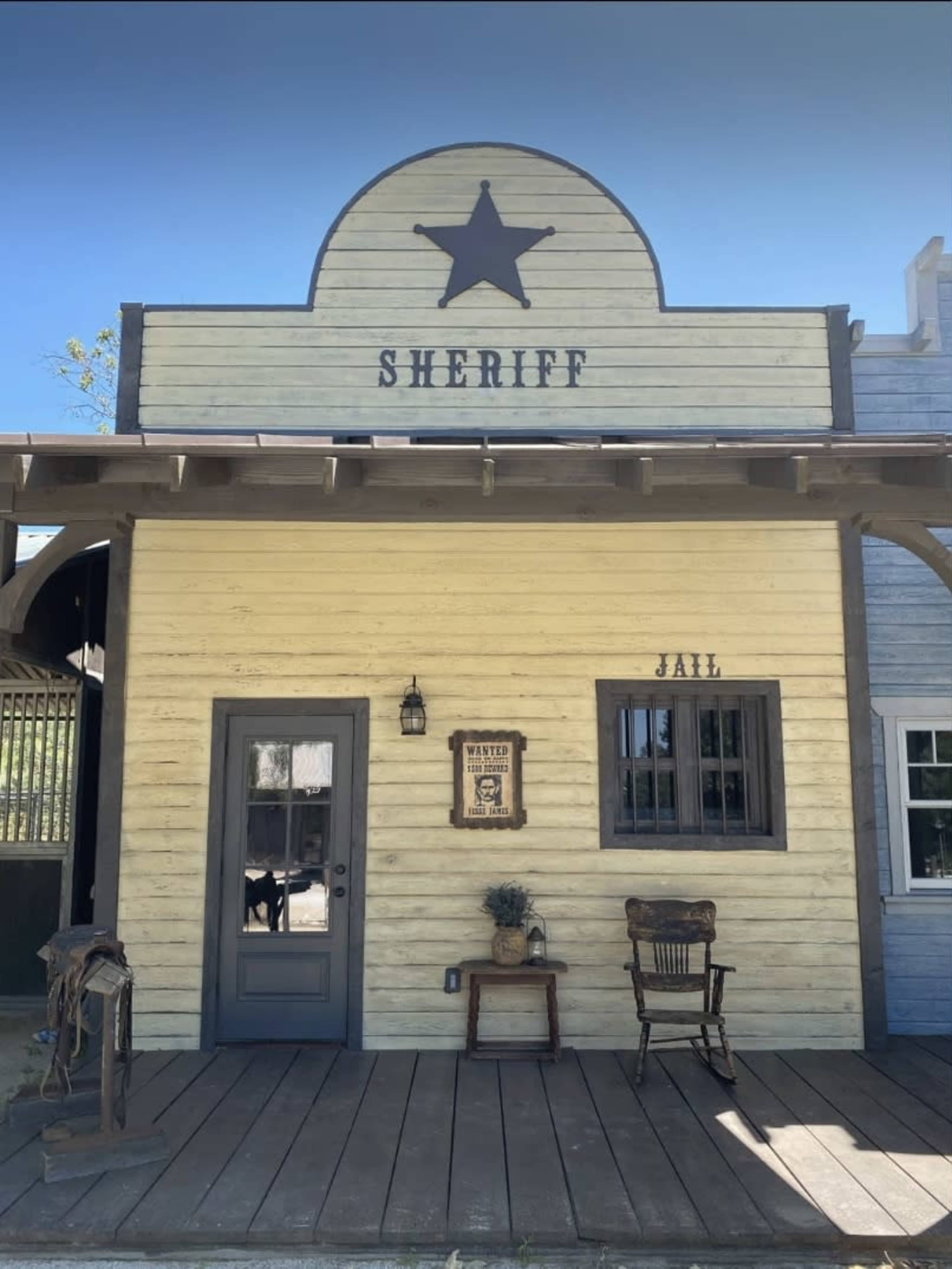 The image shows a wooden façade of a sheriff's office featuring a large star emblem, a "Jail" sign, and a wanted poster on the wall.