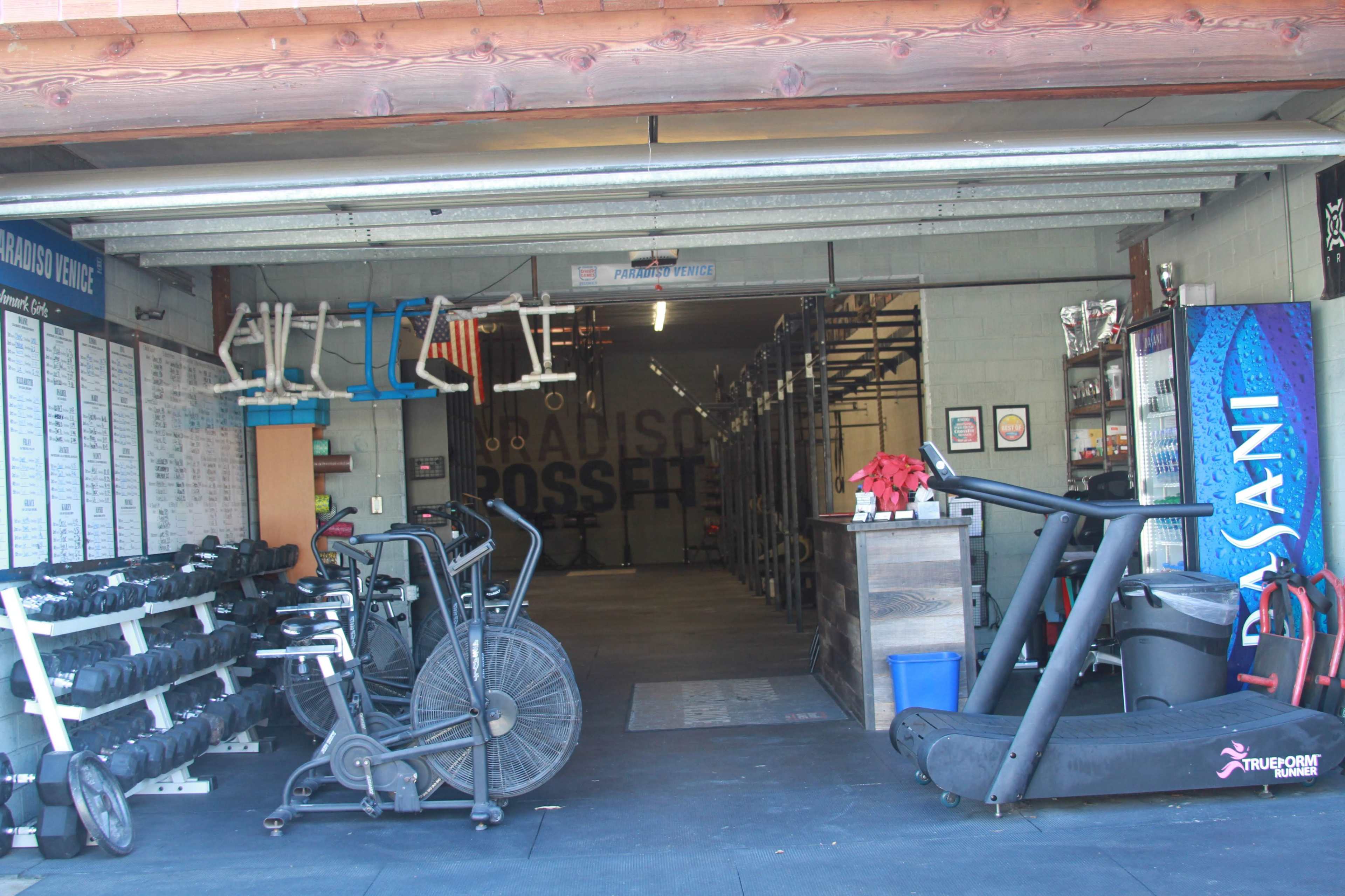 The entrance of a gym with various exercise equipment, including dumbbells, stationary bikes, and a treadmill, in a spacious, organized layout.