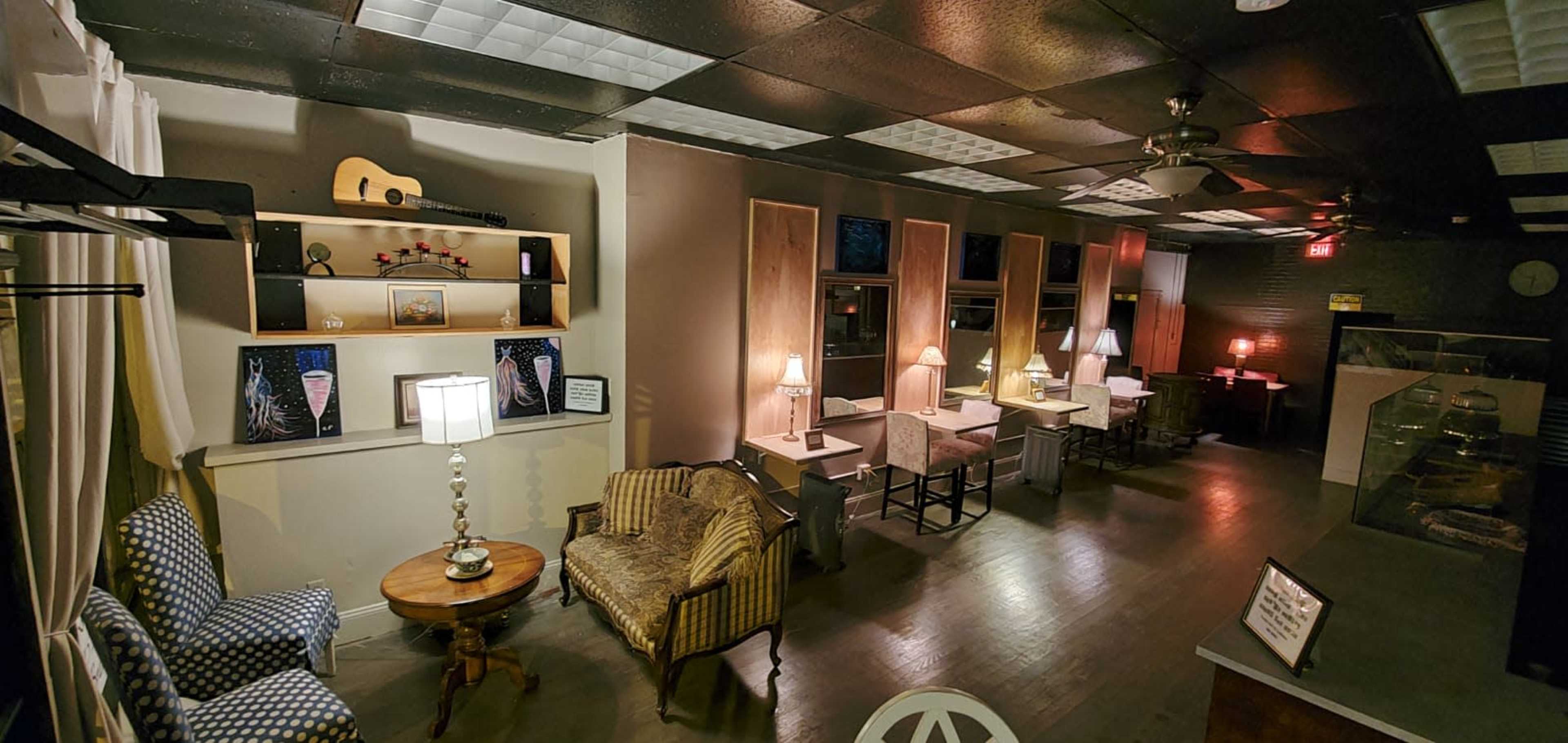 A cozy cafe interior with tables, chairs, and a small stage area, illuminated by warm lamps and accented by decorative shelves and furniture.