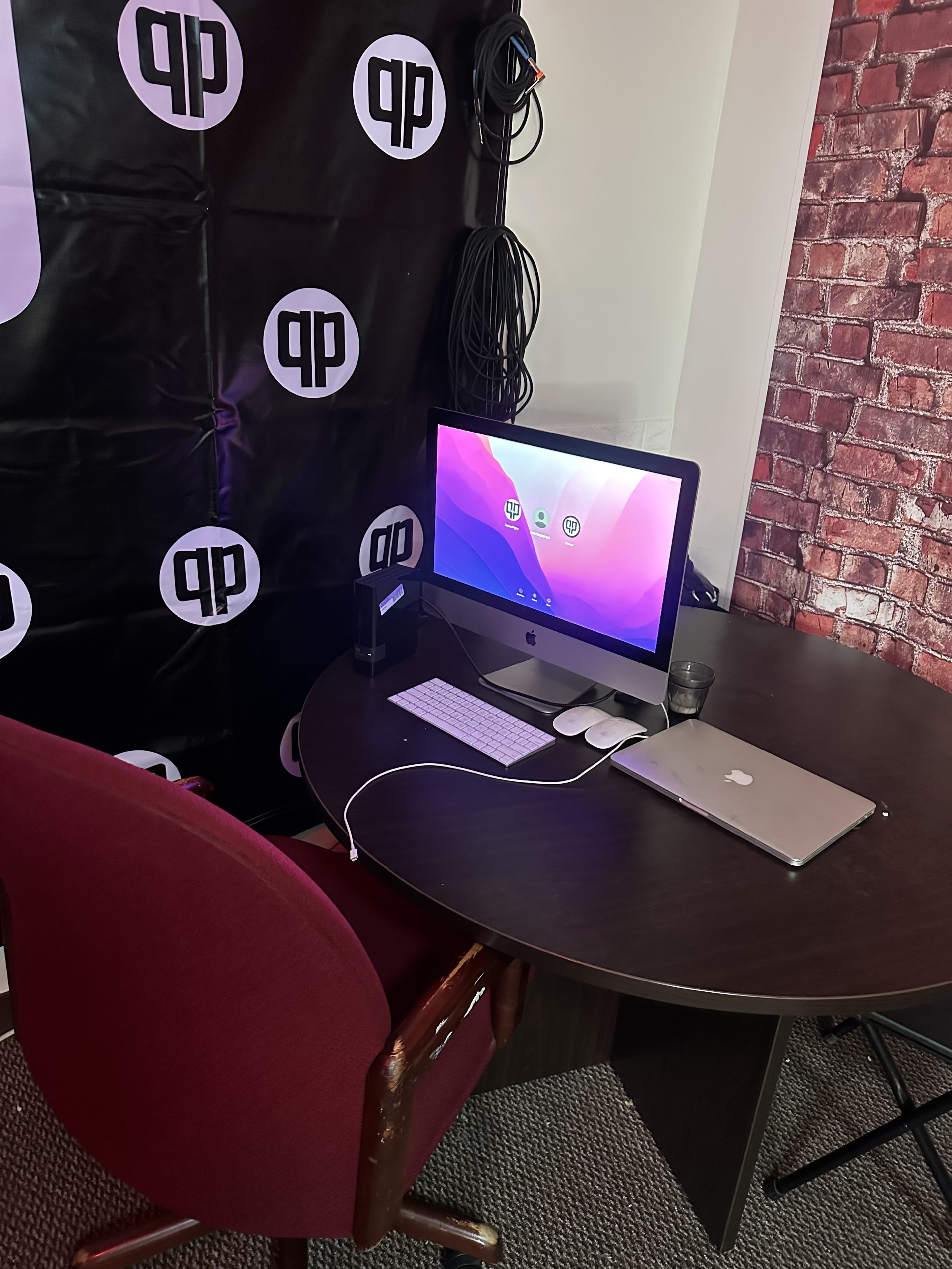 The image shows a workspace with a round table, a maroon office chair, an iMac computer, a laptop, and a decorative brick wall in the background.