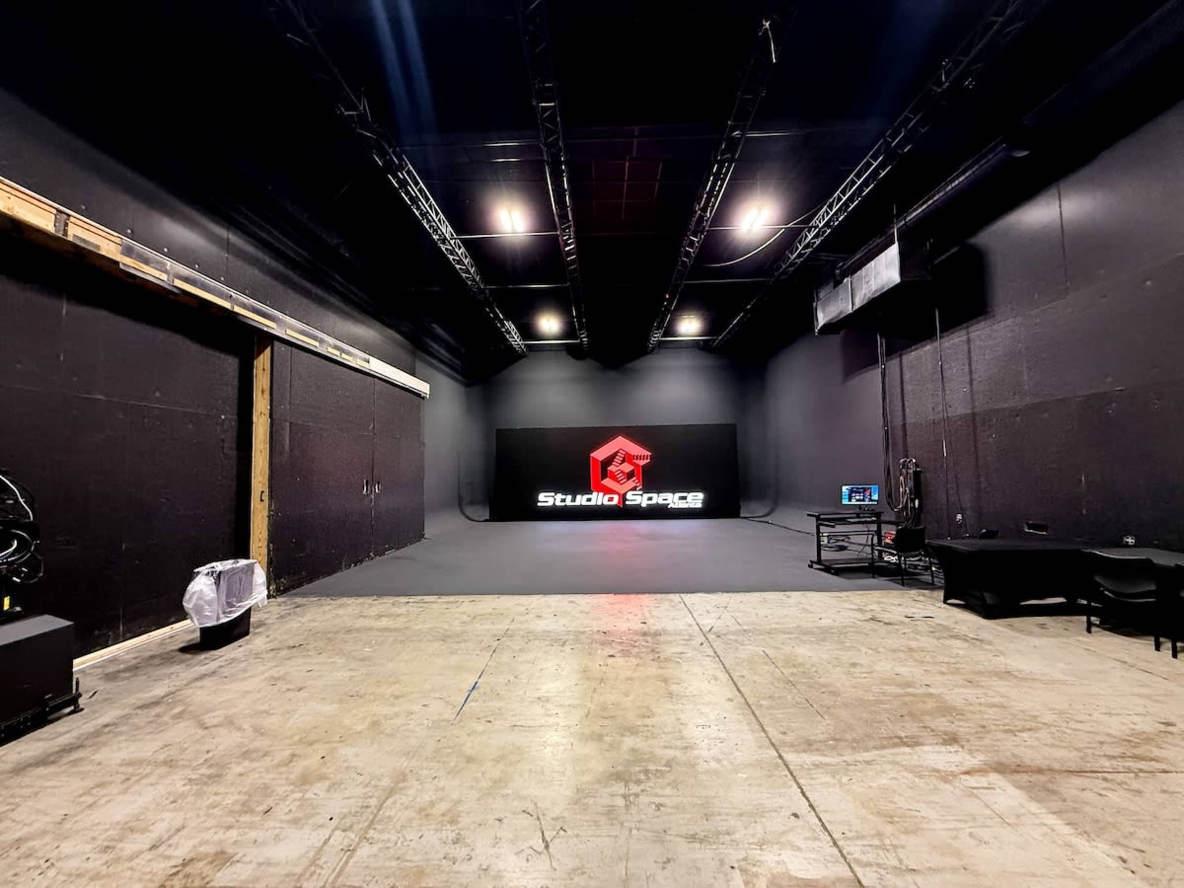 The image shows a large, dimly lit studio space featuring a black backdrop, a logo display reading "Studio Space," and various equipment along the walls.