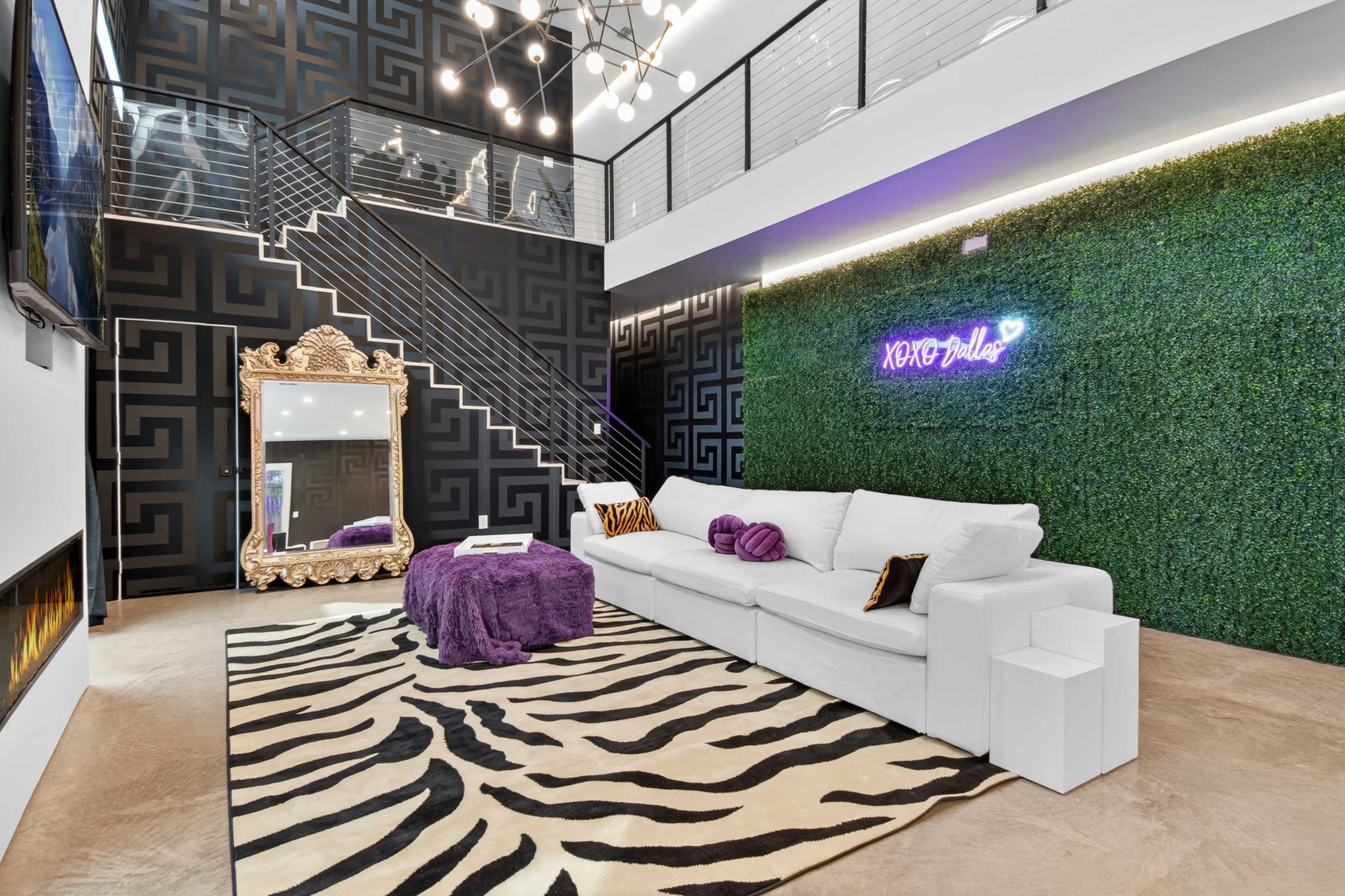 The scene features a modern living room with a white sectional sofa, a purple ottoman, a large decorative mirror, and a wall covered with artificial greenery, set against a black patterned backdrop.
