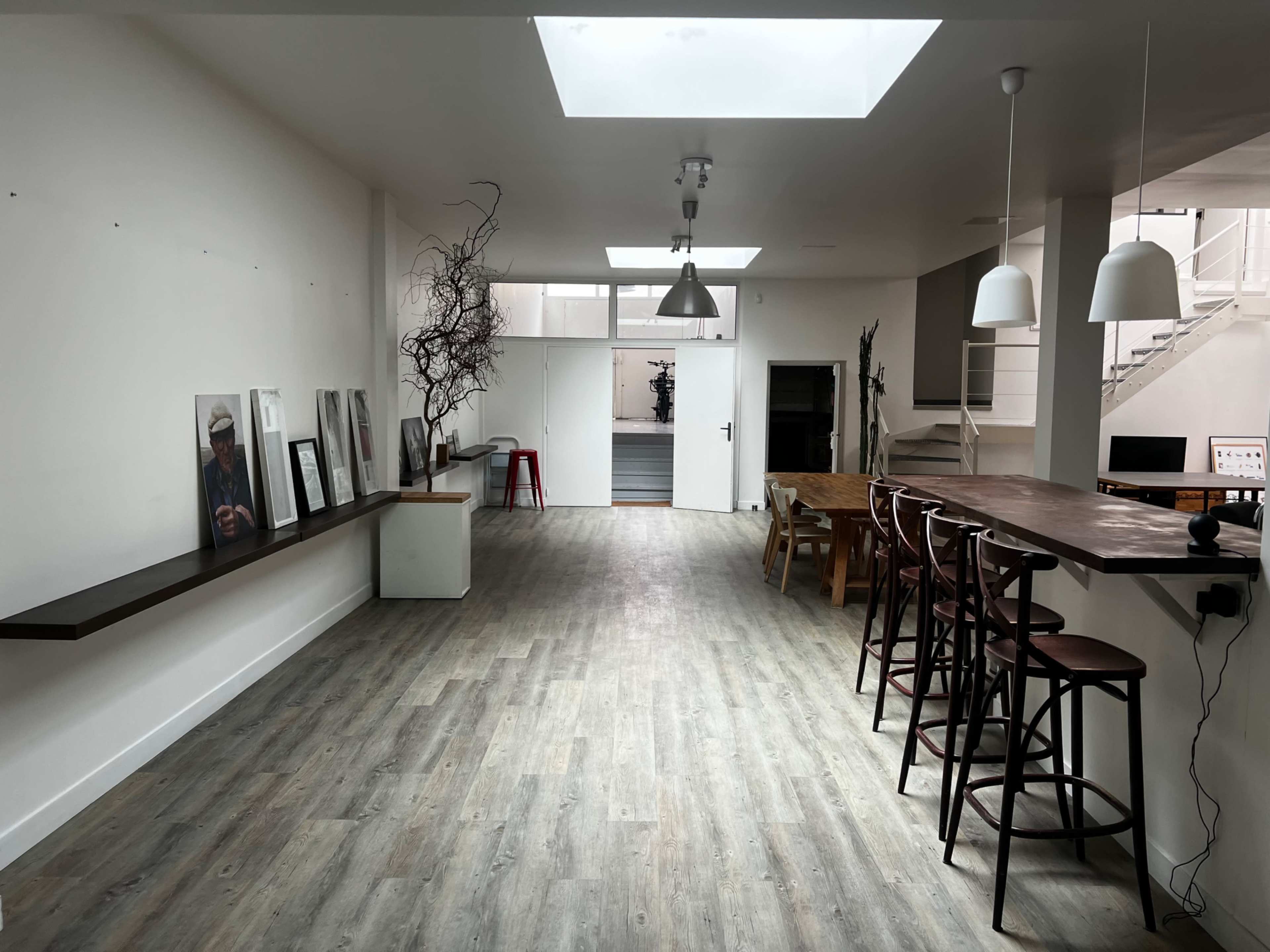 The image shows a spacious, minimalistic interior with a wooden floor, a bar-style seating area, framed pictures on the wall, and large windows letting in natural light.