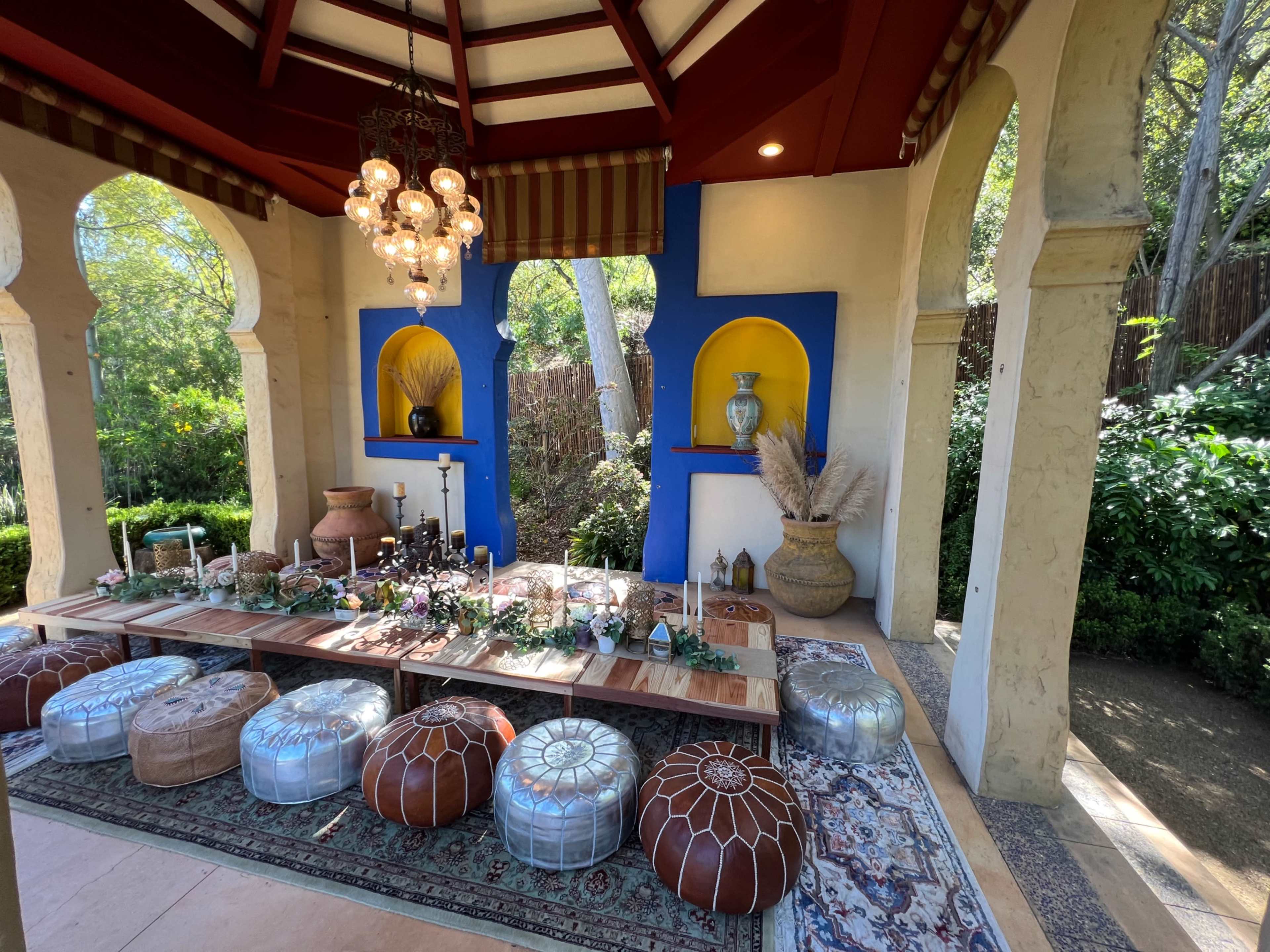 The image shows a decorative outdoor seating area with a long wooden table adorned with plants and candles, surrounded by colorful poufs and a vibrant background.