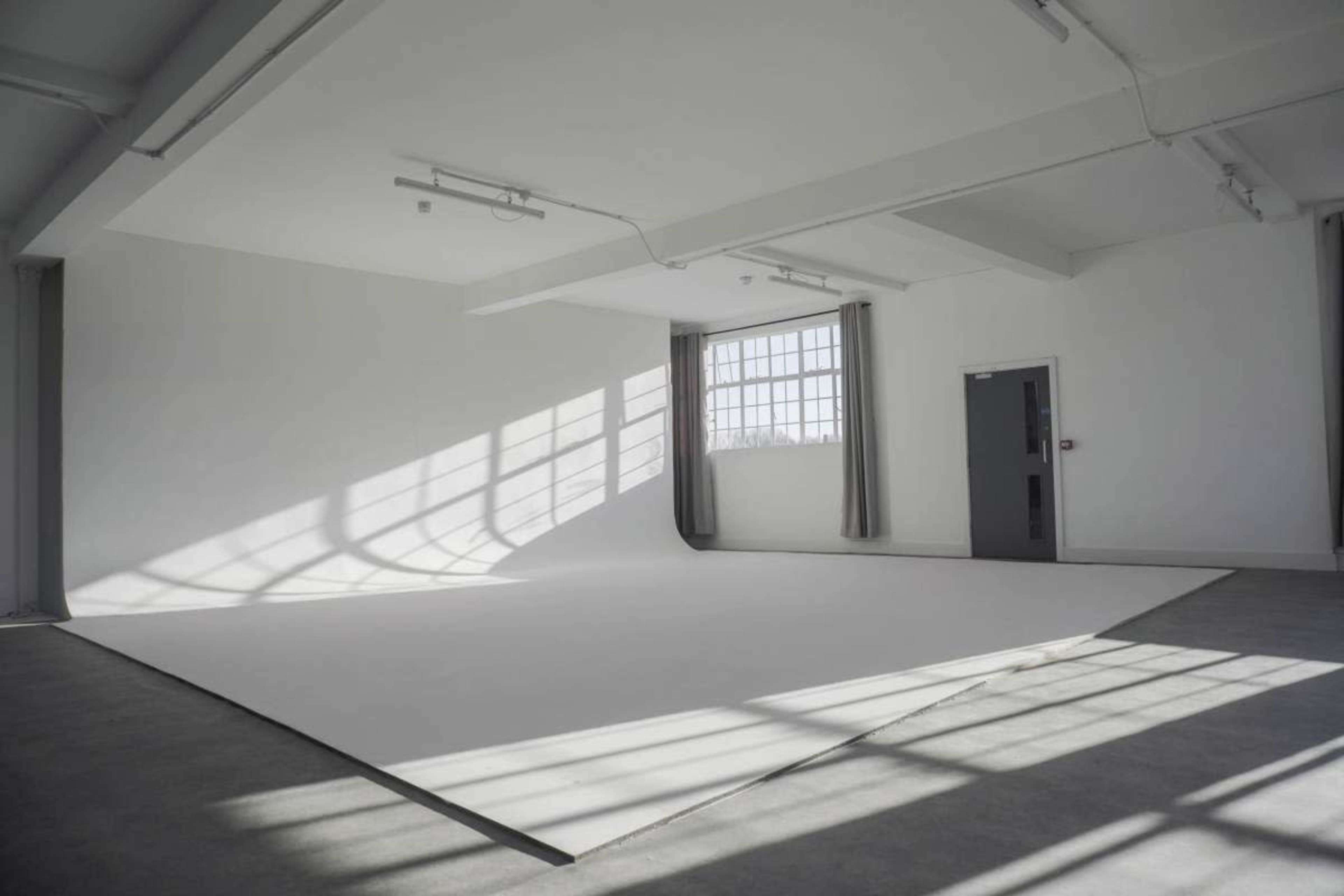 The image shows a large, empty studio space with a white backdrop and natural light streaming through a window, casting shadows on the floor.