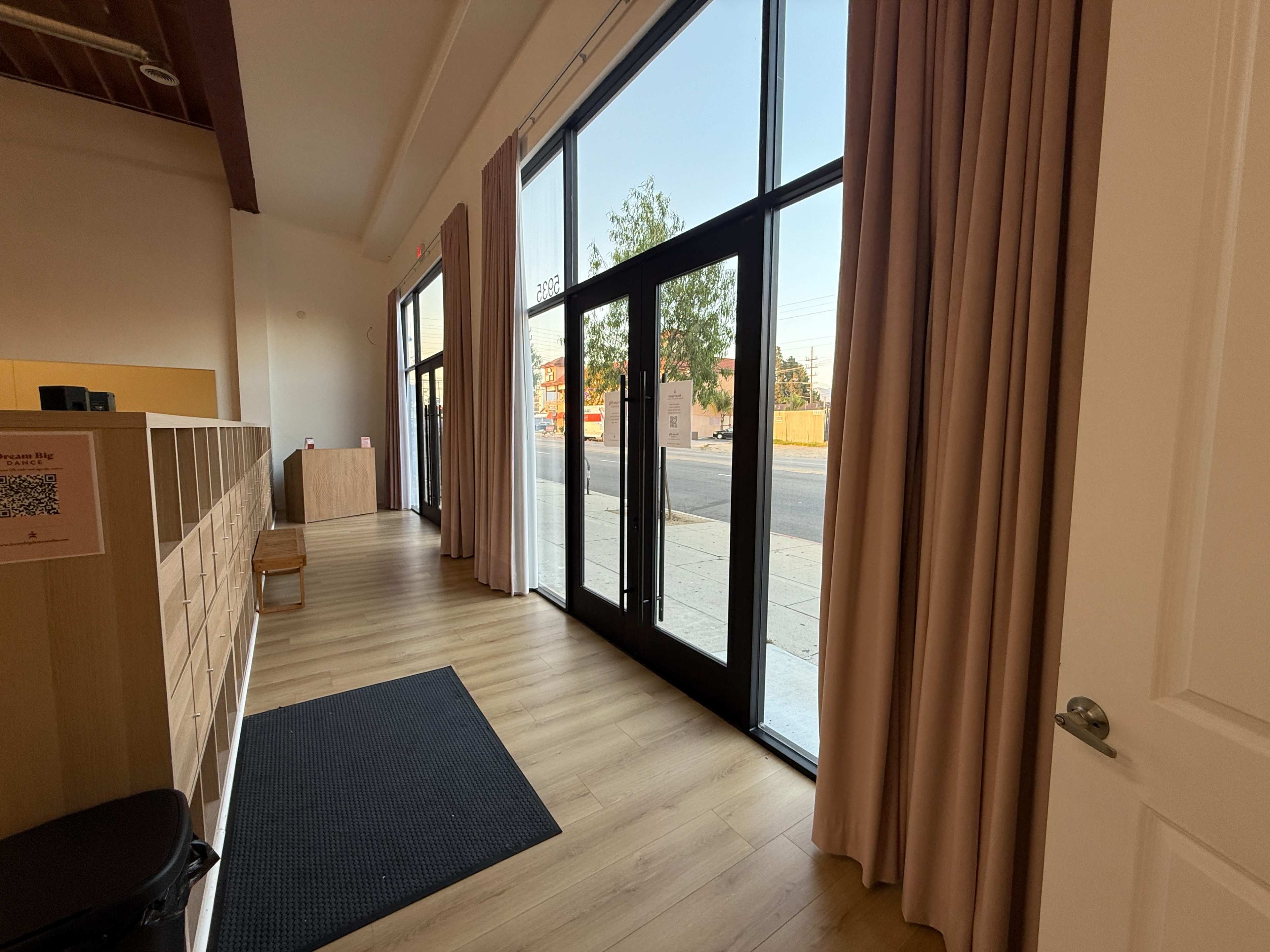 The image shows a modern interior entrance with large glass doors, wooden flooring, and minimalistic furniture.