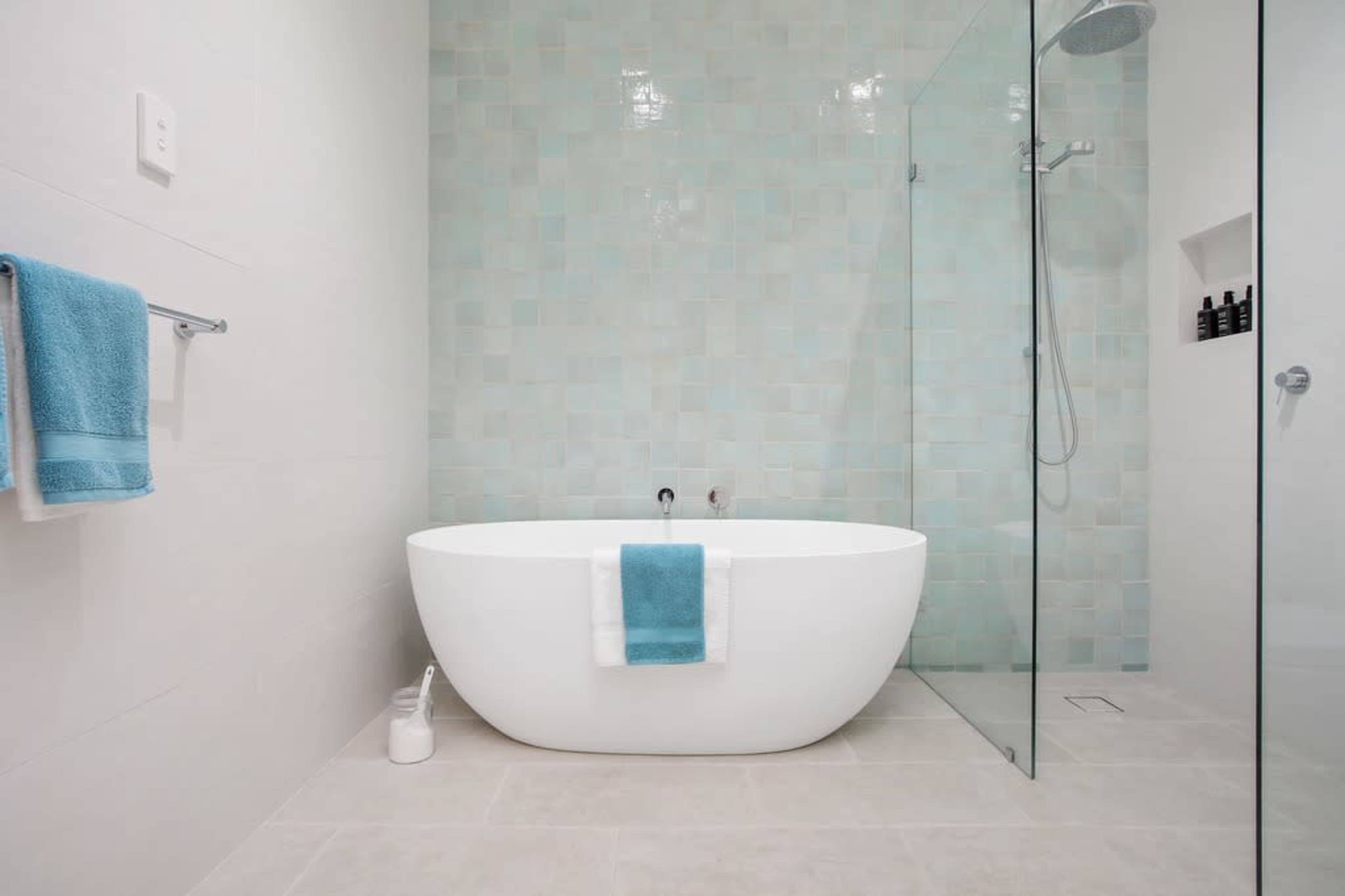 A modern bathroom featuring a freestanding white bathtub with a turquoise towel, glass shower enclosure, and light-colored tiled walls.