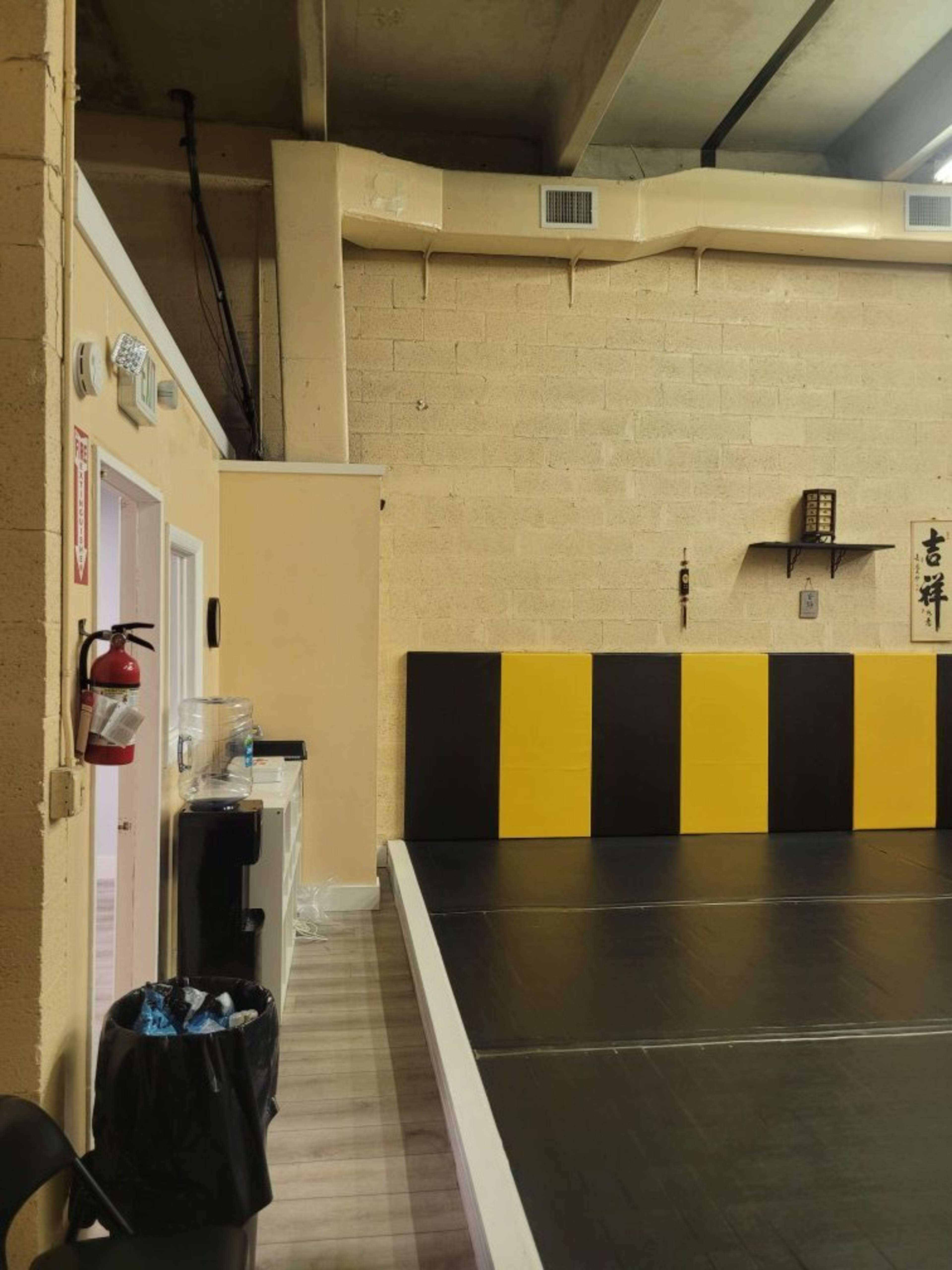 The image shows a corner of a martial arts studio with a black and yellow padded wall, a garbage can, a fire extinguisher, and a doorway leading to another room.
