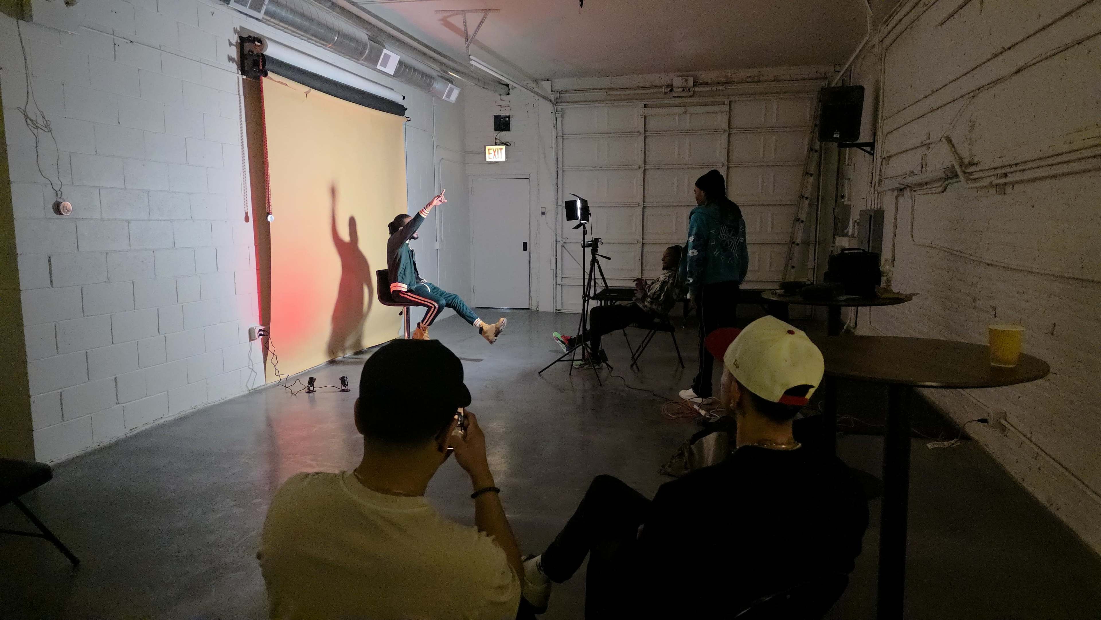 A person in a teal outfit poses on a stool against a yellow backdrop while shadows are cast on the wall in a studio setting.