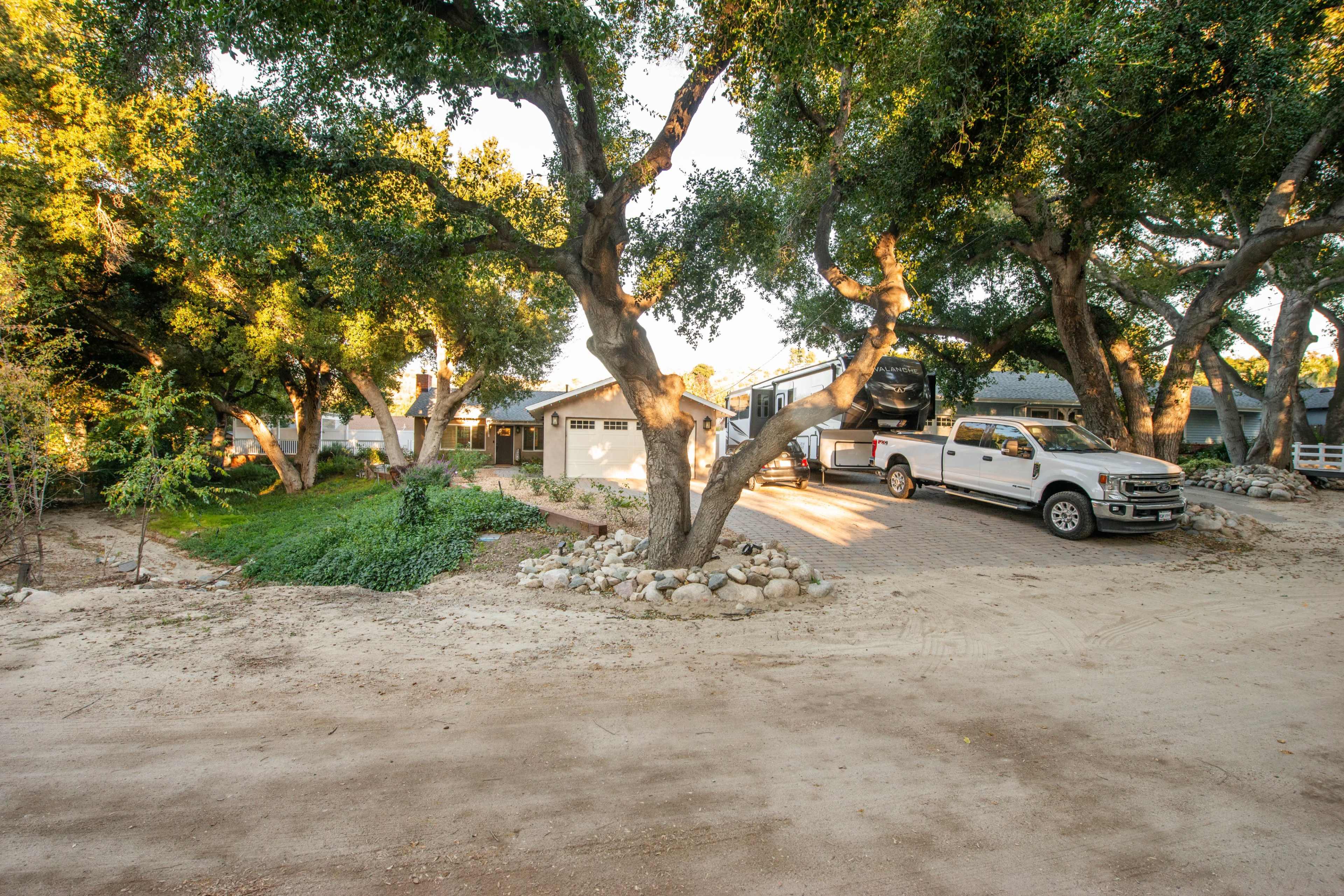 A white truck is parked in a driveway next to a house surrounded by trees and gravel.