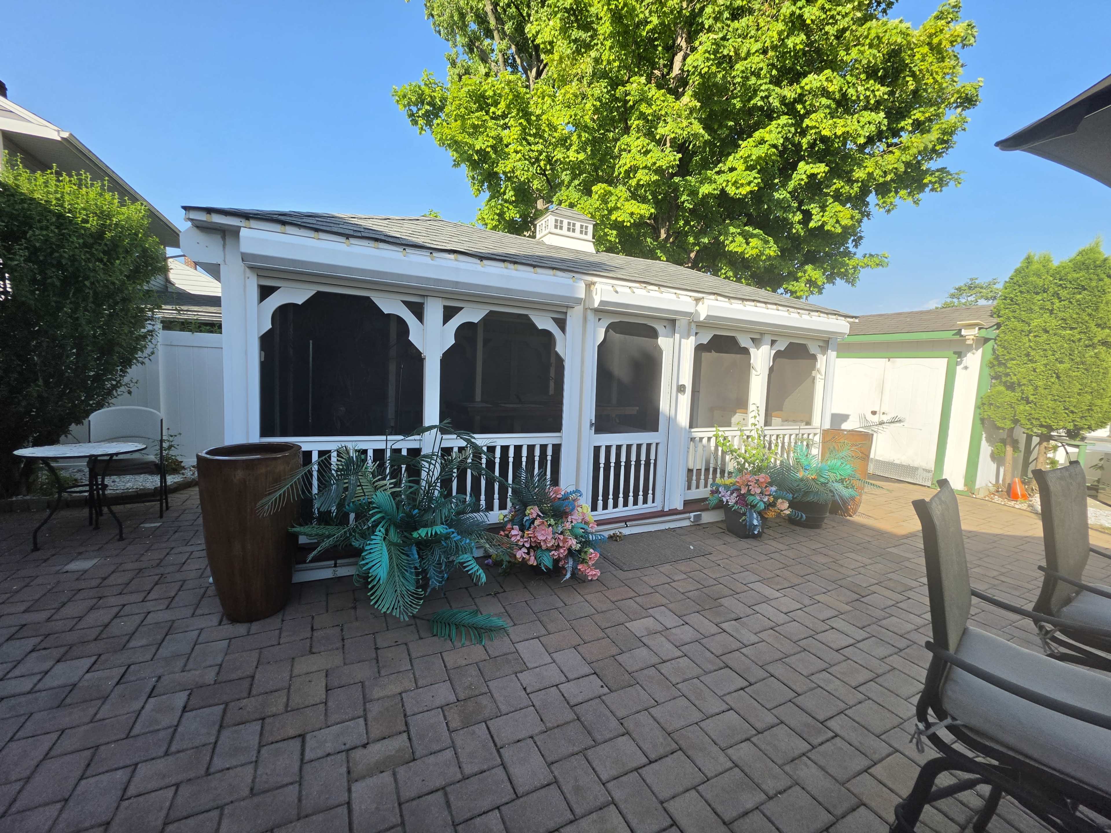 The image shows a white gazebo-style structure in a landscaped backyard with potted plants and a brick patio.