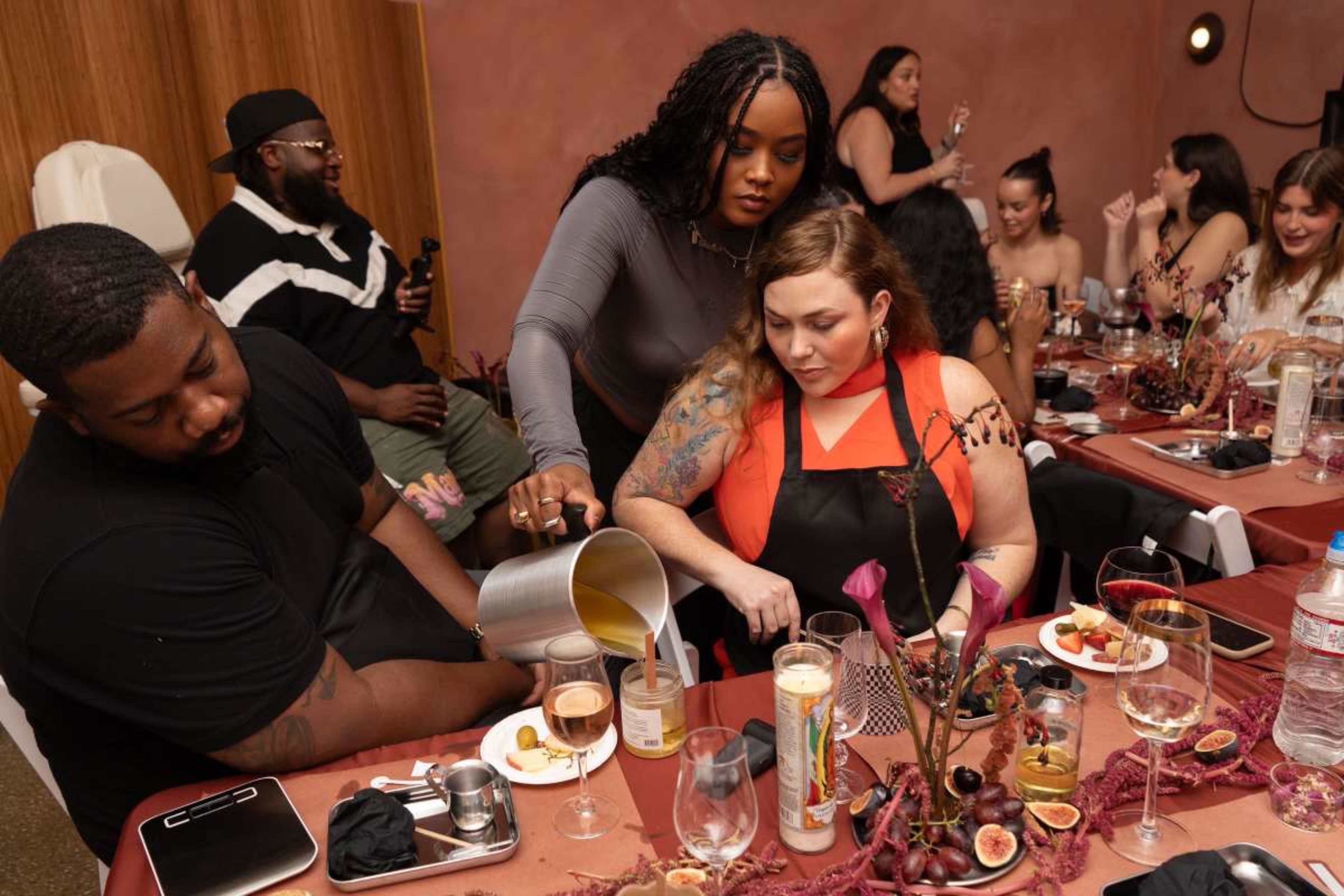 A group of people is seated around tables in a warmly decorated dining area, while one woman in an apron pours a drink for another woman.