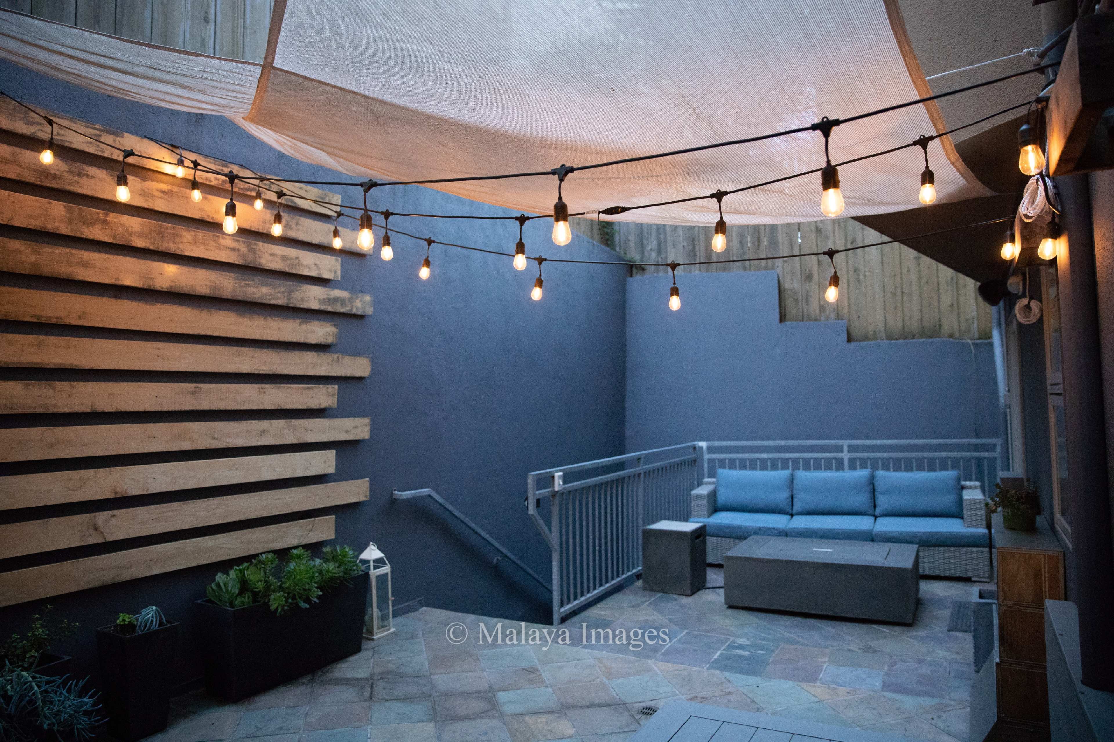 The image shows a cozy outdoor space with string lights, a seating area, and wooden wall accents.