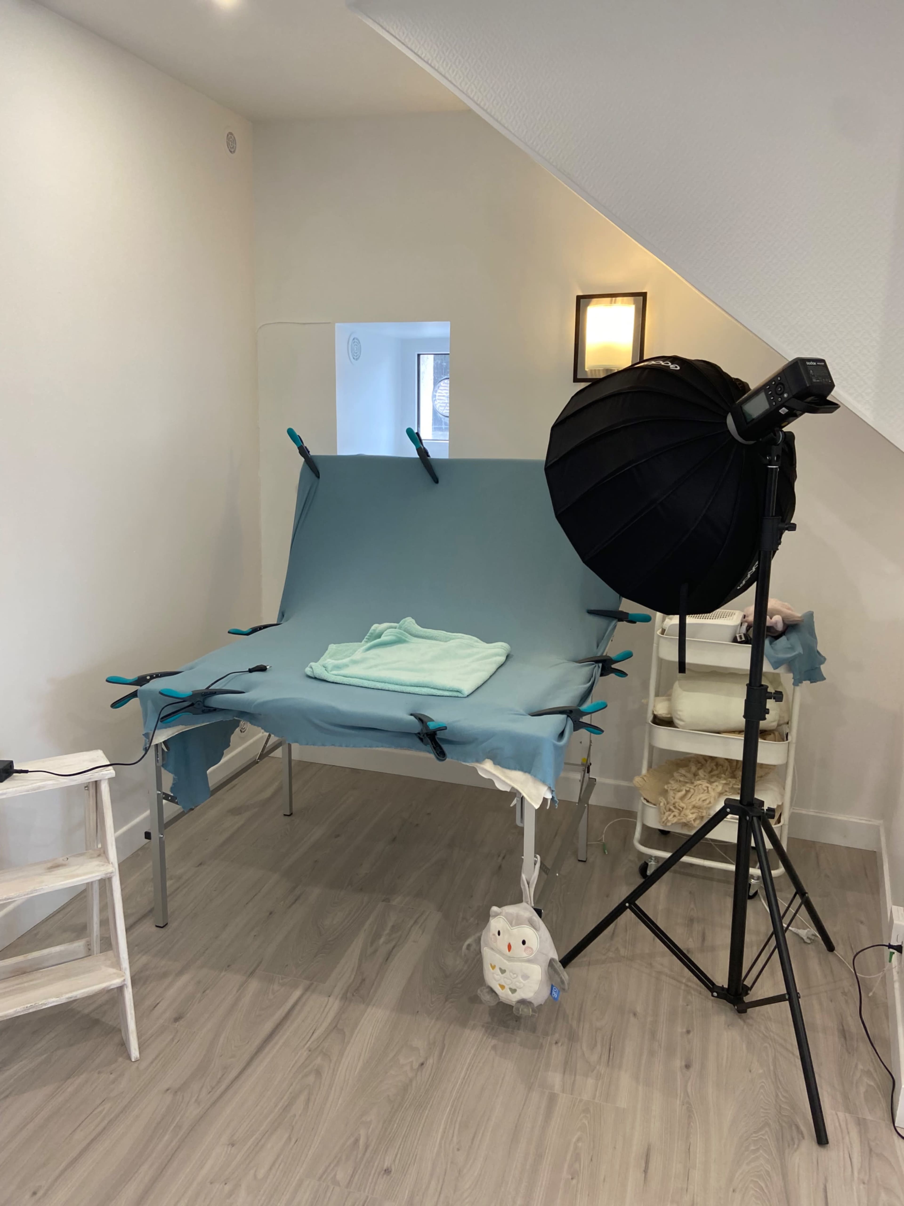 A small studio setup with a blue backdrop draped over a folding table, a photography light on a stand, and a shelf with materials in the corner.