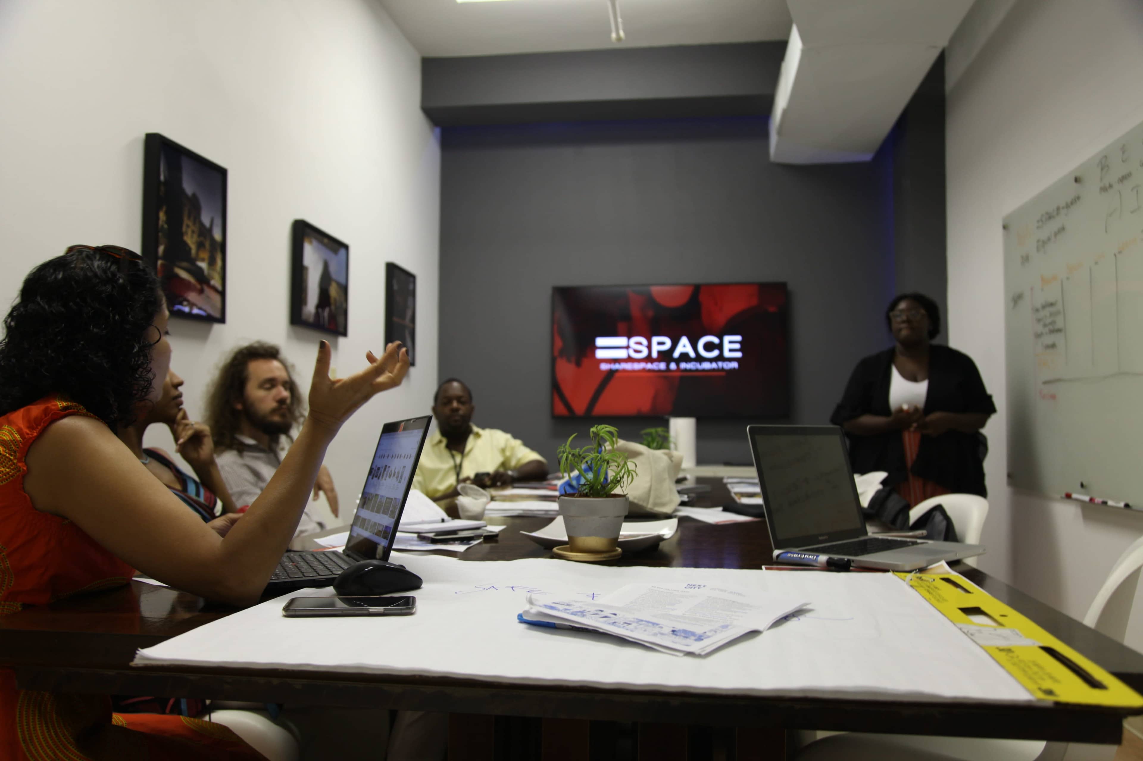 A group of people is seated around a conference table in a meeting room, discussing while using laptops and notes, with a television screen displaying the word "SPACE" on the wall.