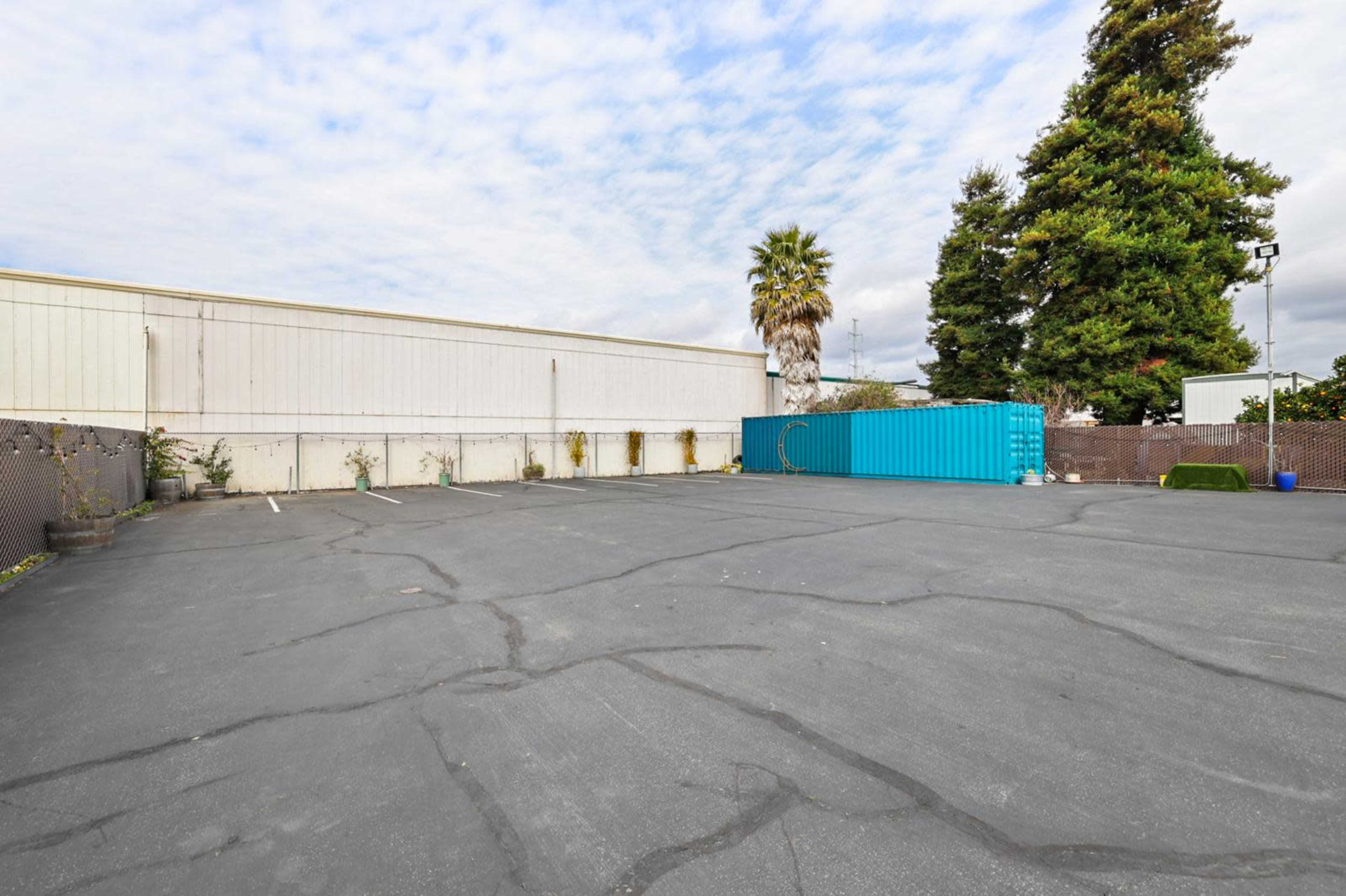 The image shows an empty asphalt parking lot with a blue shipping container against a light-colored building and a palm tree nearby.