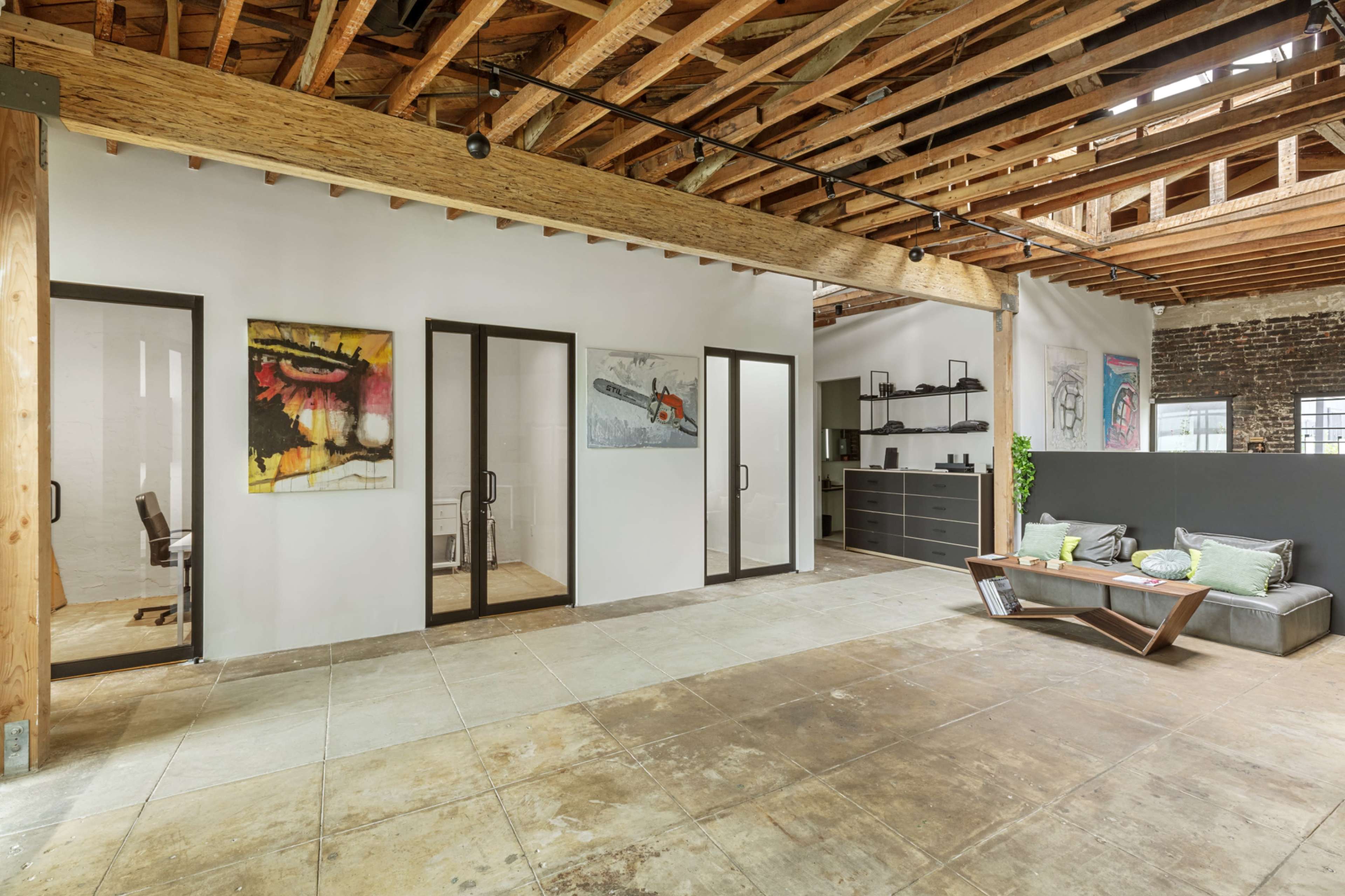The image shows a spacious open office area with exposed wooden beams, several artworks on the walls, and glass-enclosed offices.