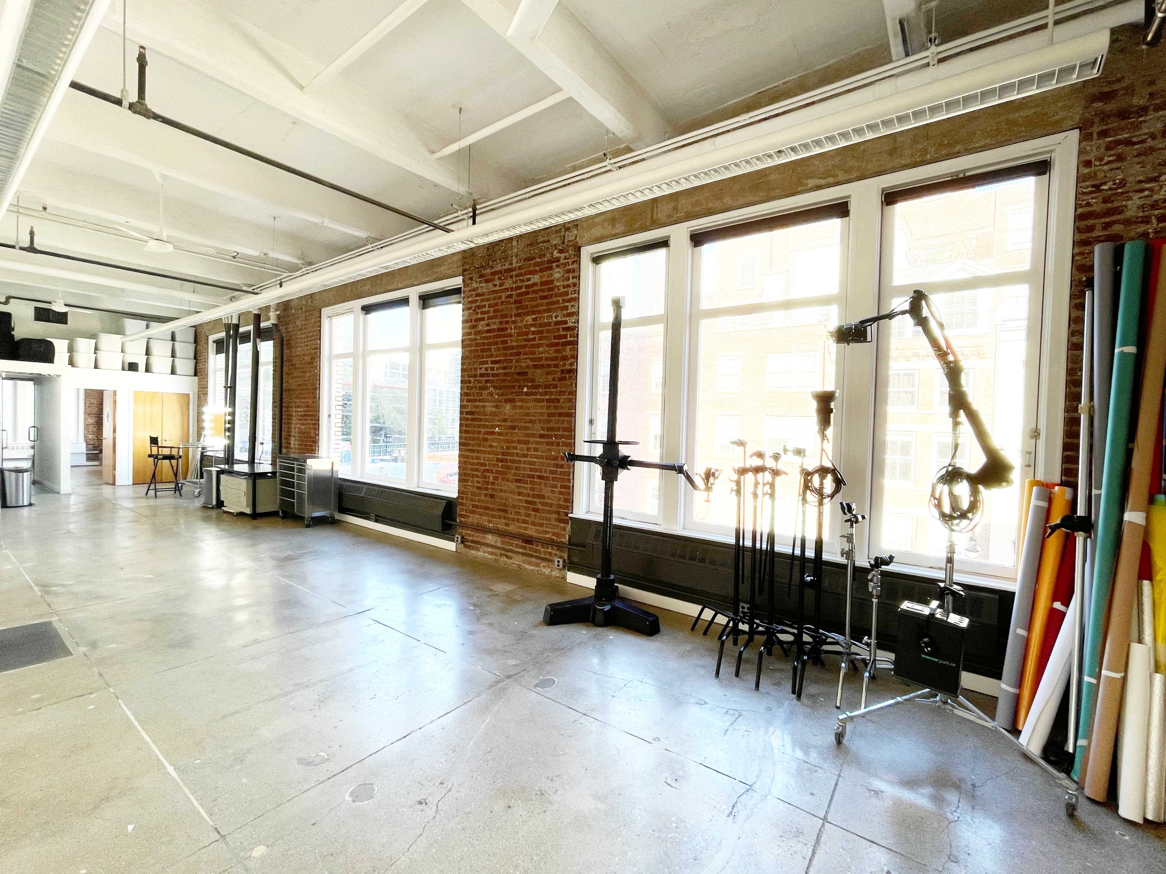 A spacious studio features large windows, exposed brick walls, and equipment stands along one side.