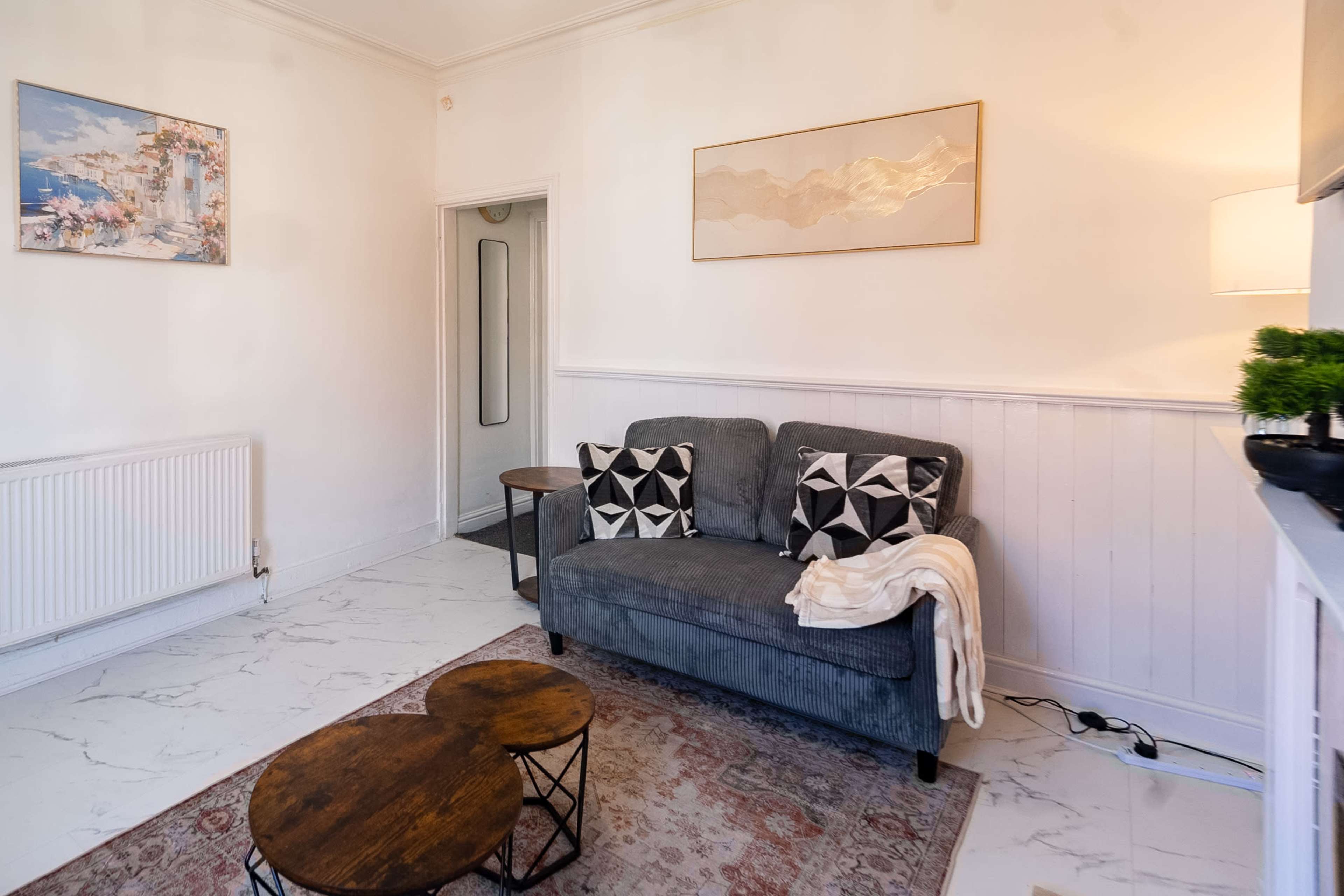 The image shows a cozy living room with a gray sofa, two decorative pillows, a wooden coffee table, and a wall-mounted art piece.