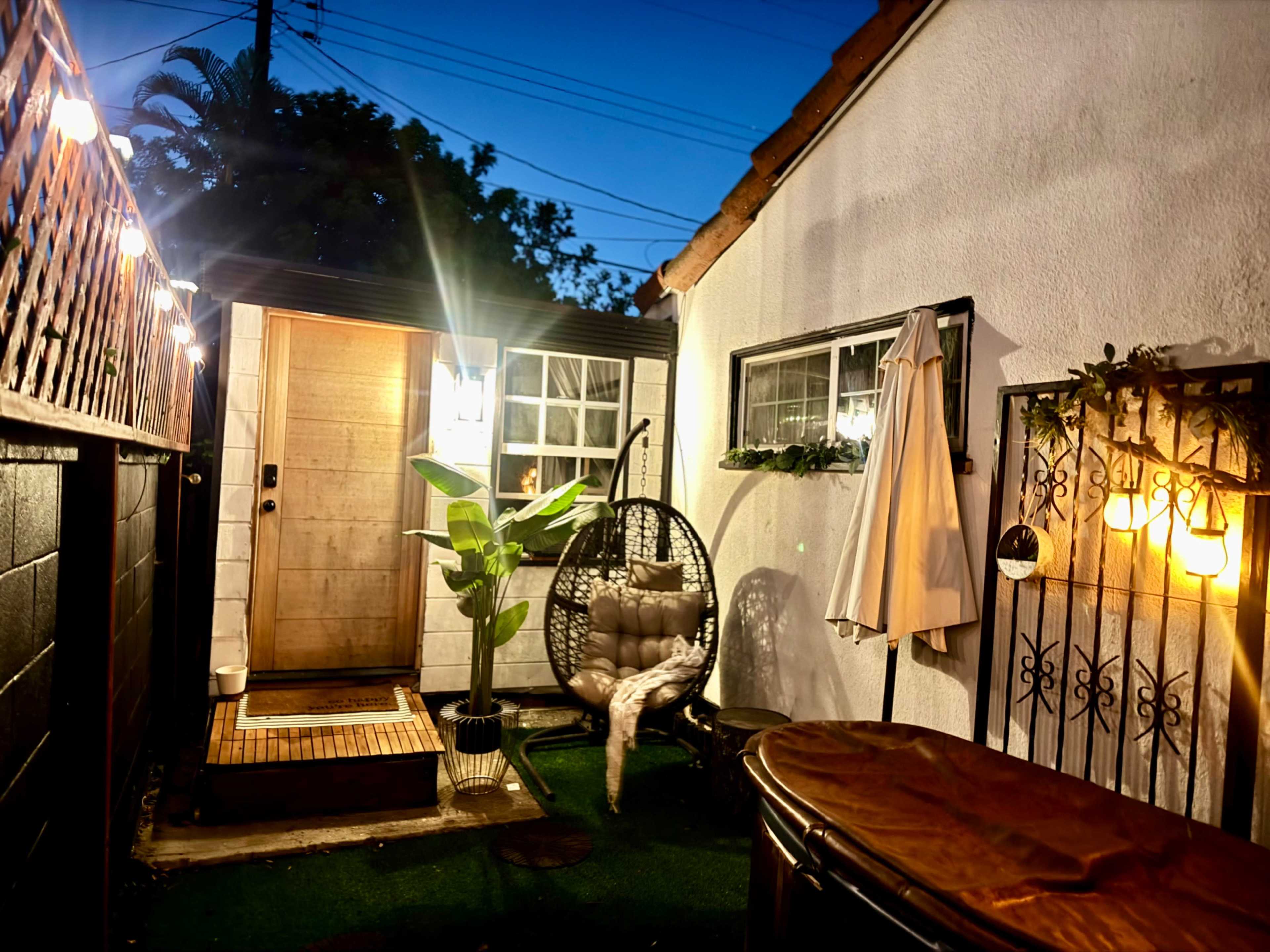 A cozy outdoor space features a swinging chair, a hot tub, and a small entrance, all illuminated by string lights against a twilight sky.