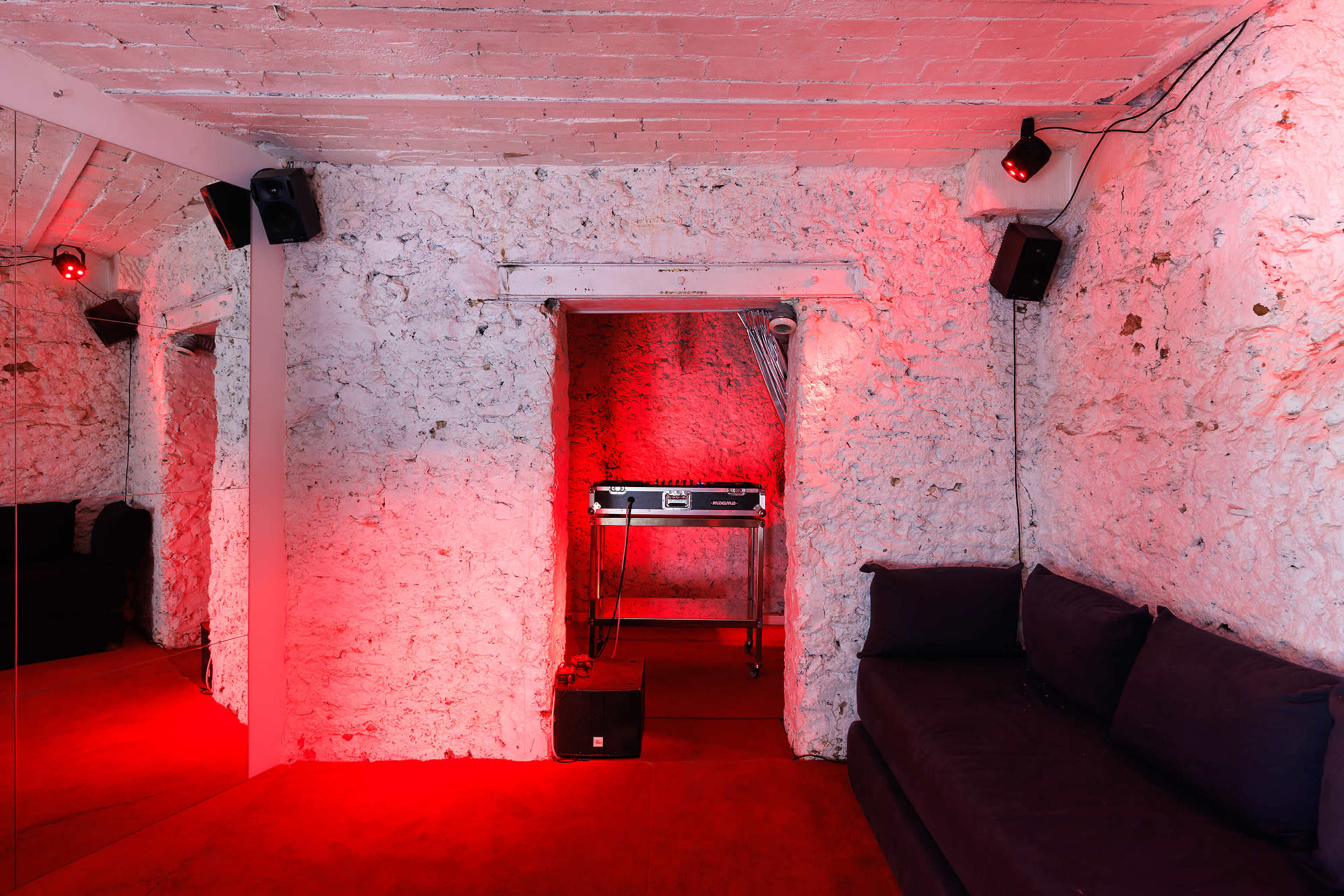 A dimly lit room with rough, whitewashed walls features a DJ setup at the back and a dark sofa along one side.
