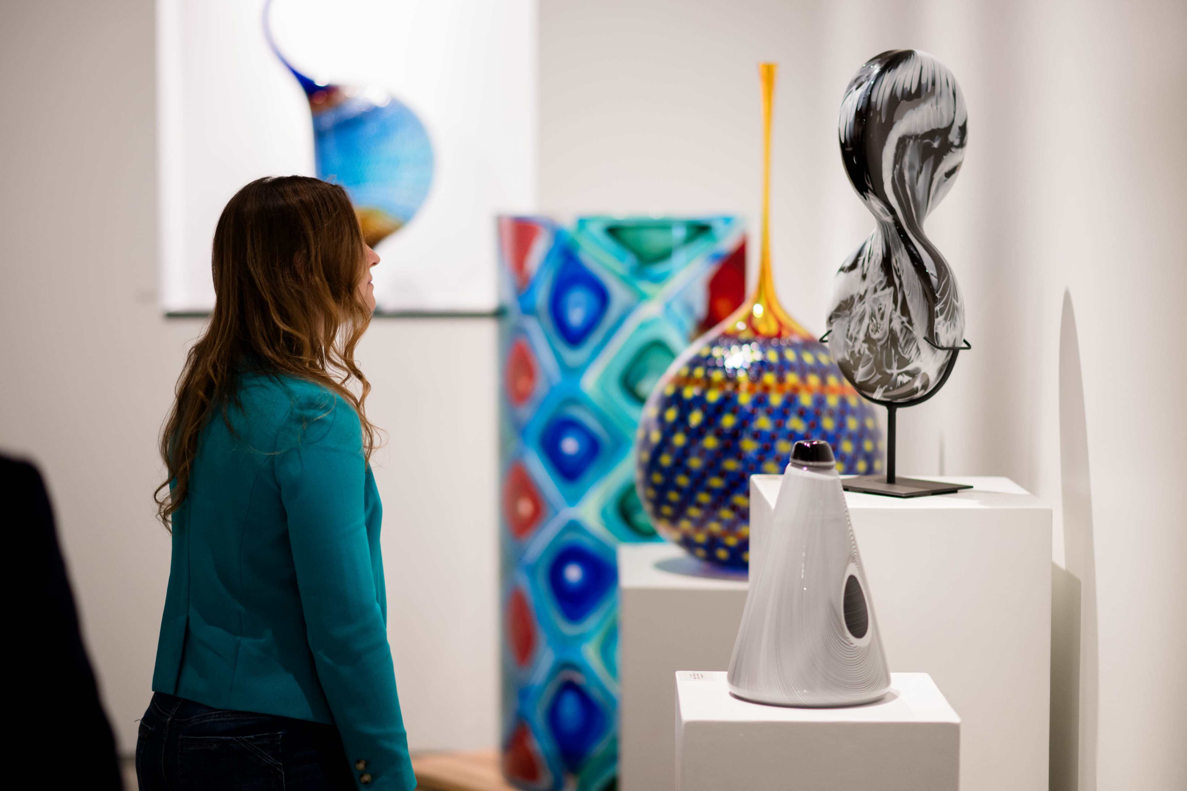 A woman in a teal jacket examines various pieces of colorful glass art displayed on white pedestals in a gallery setting.