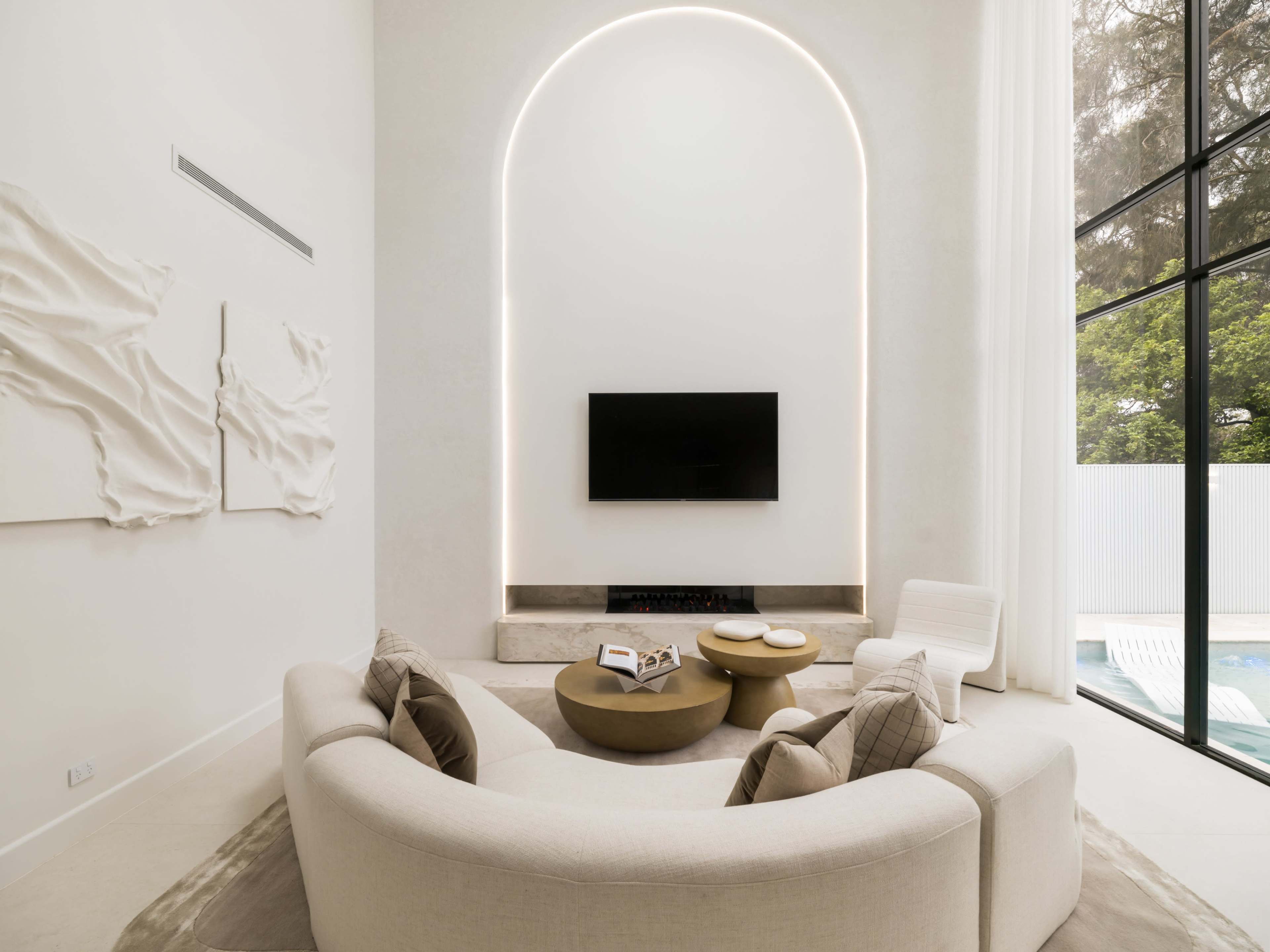 The image shows a modern living room with a curved sofa, minimalist art pieces on the wall, and a television mounted above a stone fireplace, all illuminated by soft lighting.