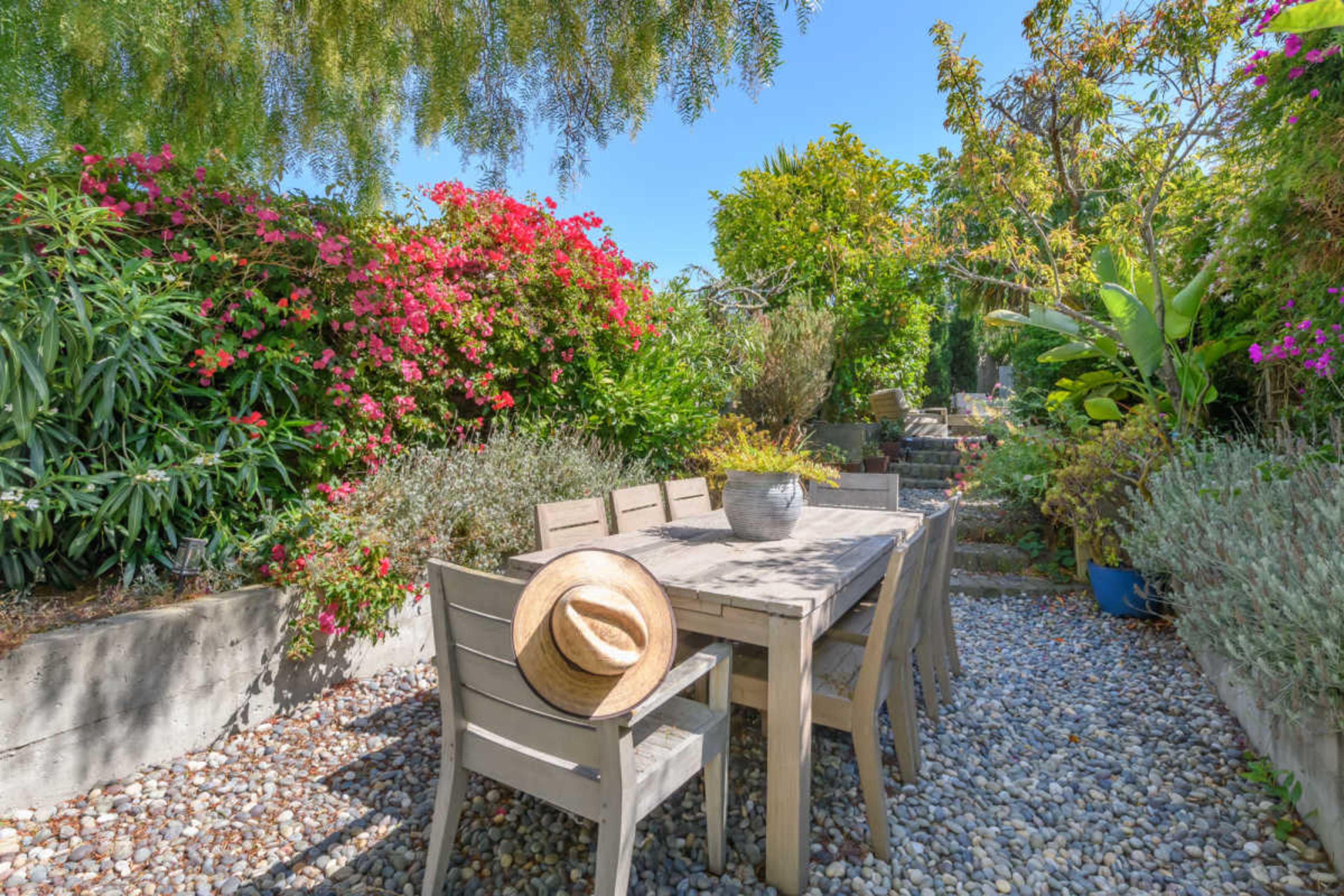 A wooden dining table with chairs is surrounded by various flowering plants and greenery in a sunny garden setting.