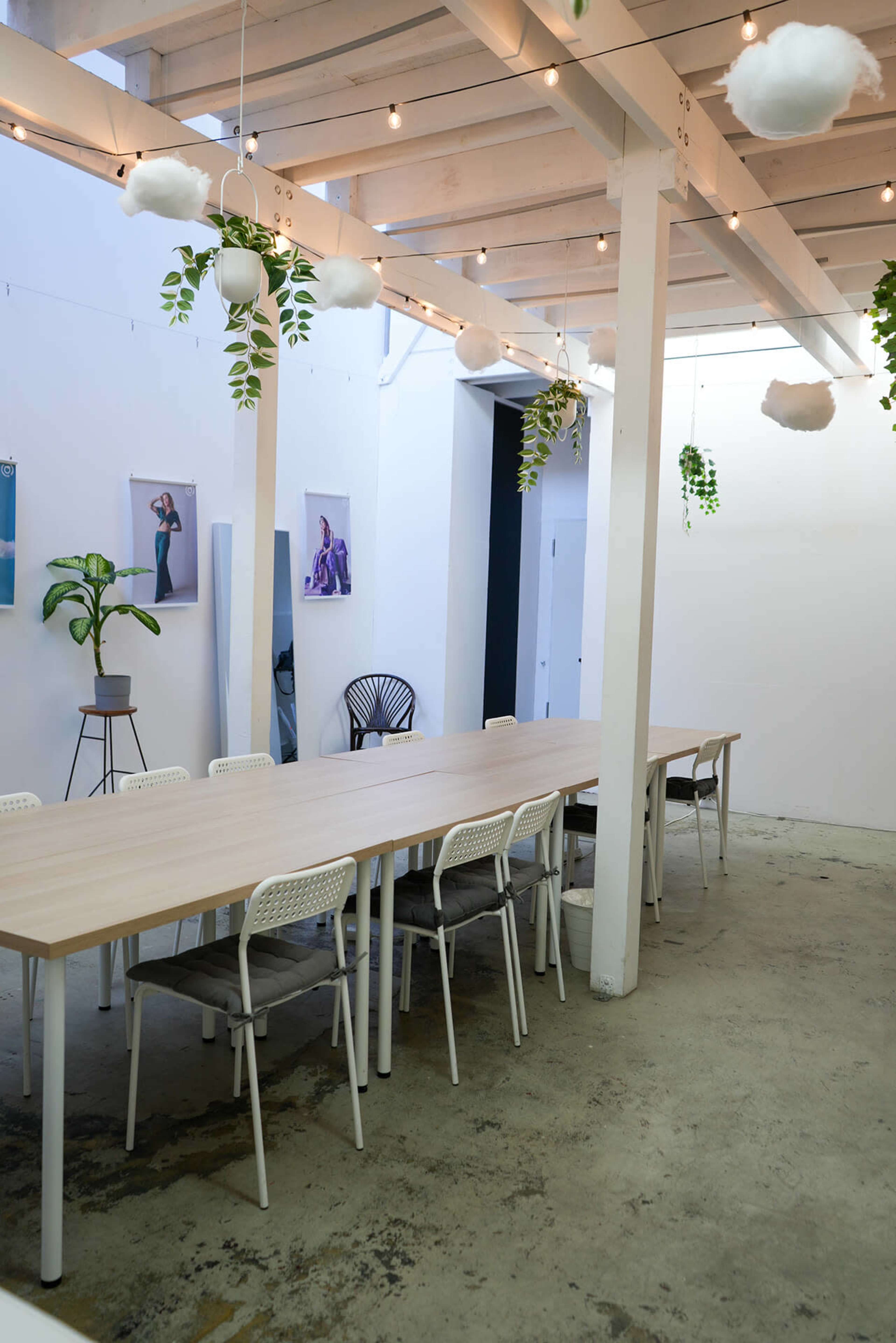 A spacious meeting room features a long wooden table surrounded by white chairs, with hanging plants and decorations suspended from the ceiling.