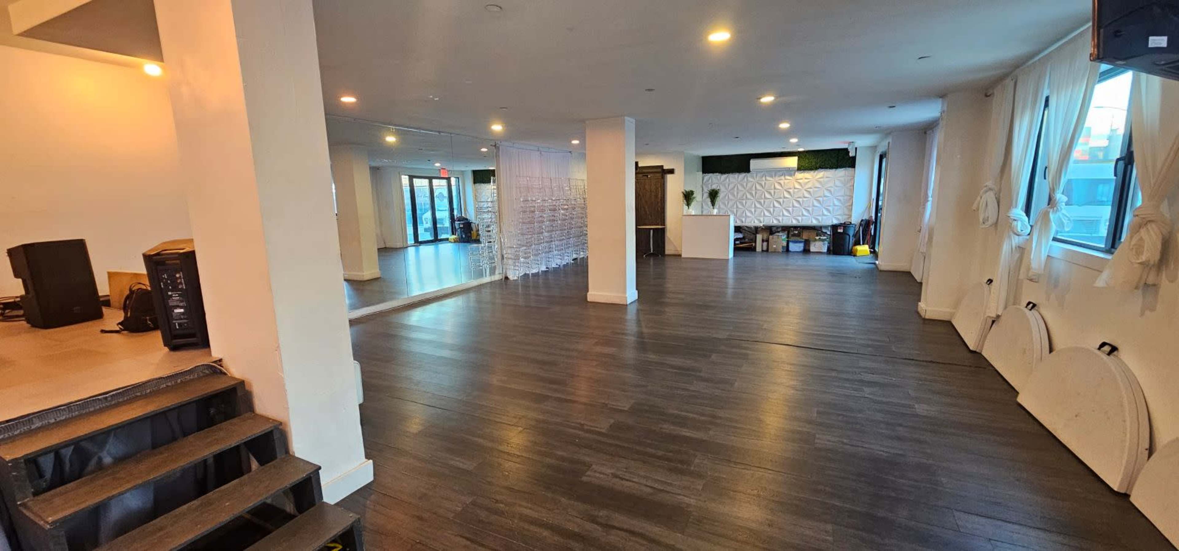 The image shows a spacious, well-lit interior room with hardwood floors, white walls, and a large mirror along one side, featuring a reception area and scattered cleaning equipment.