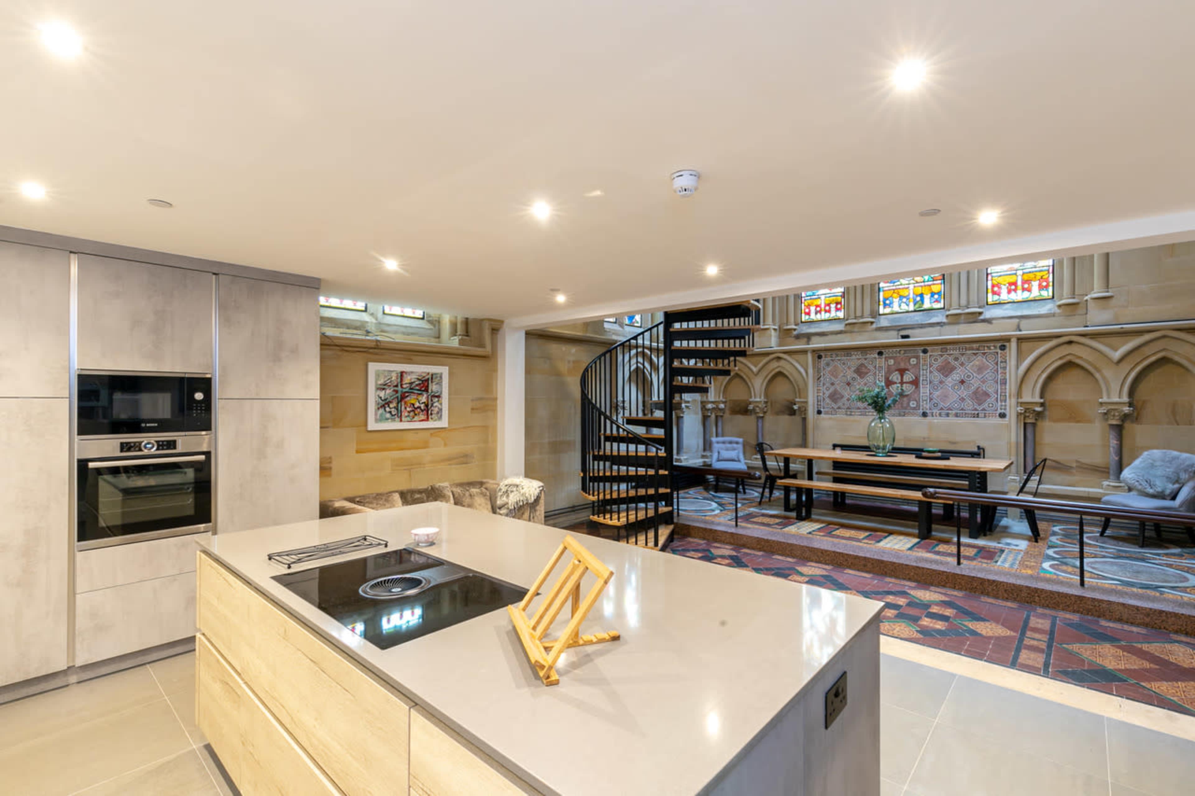 A modern kitchen with an island, featuring a spiral staircase in the background and stained glass windows illuminating the space.