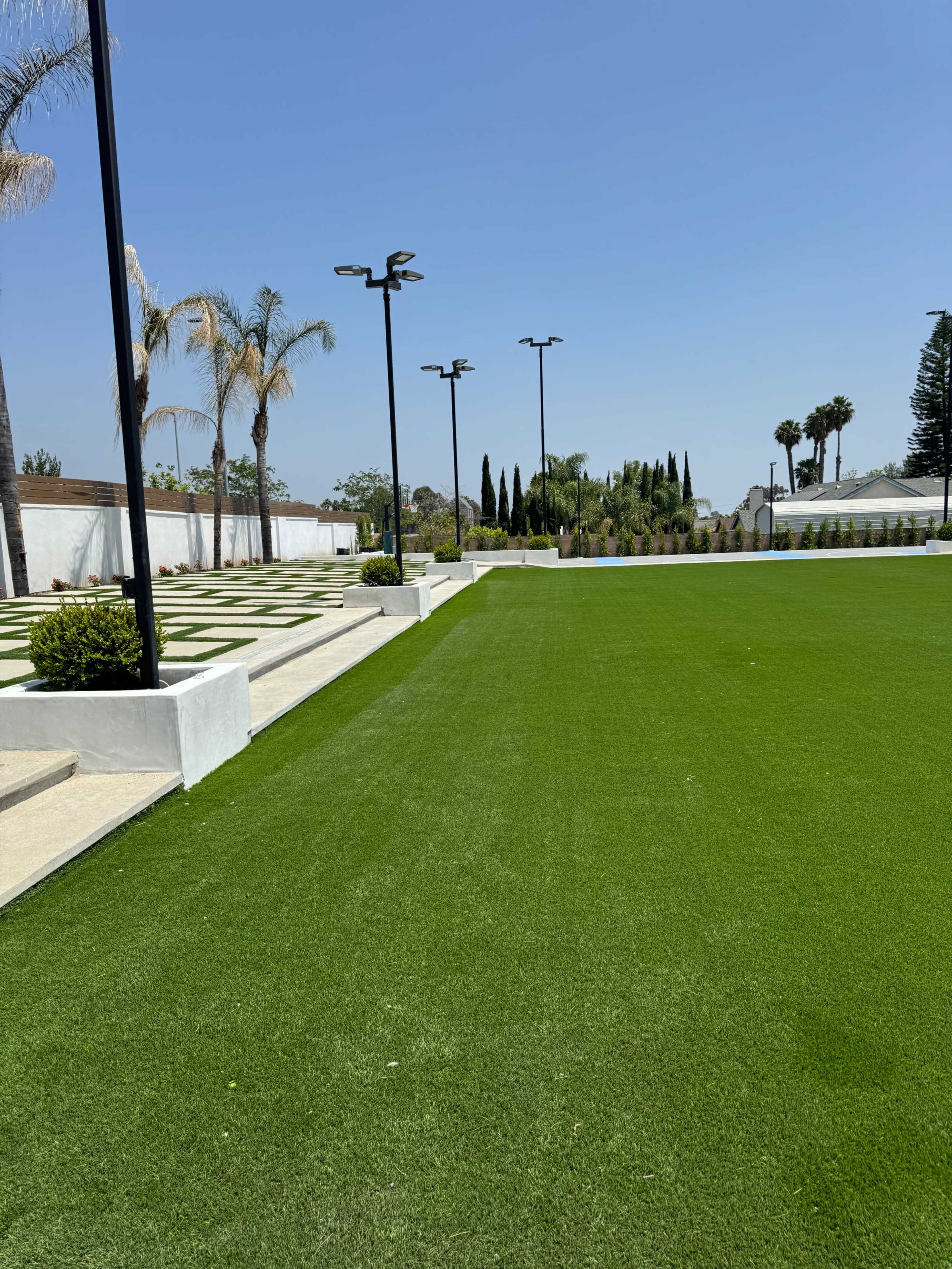 A spacious, well-maintained artificial grass area bordered by neatly arranged pathways and palm trees under a clear blue sky.