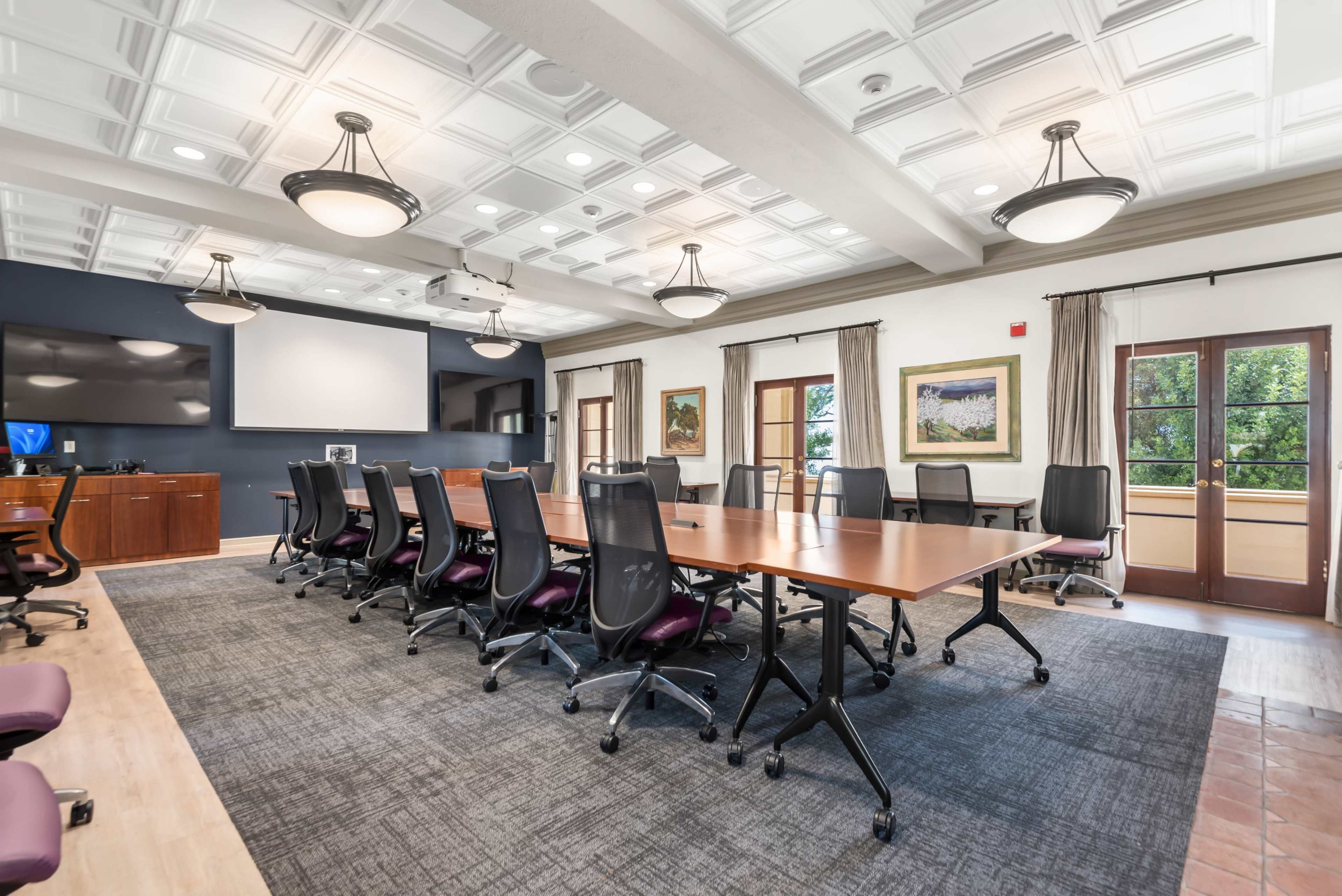 The image shows a modern conference room equipped with a large wooden table, ergonomic chairs, and multiple windows allowing natural light to enter.