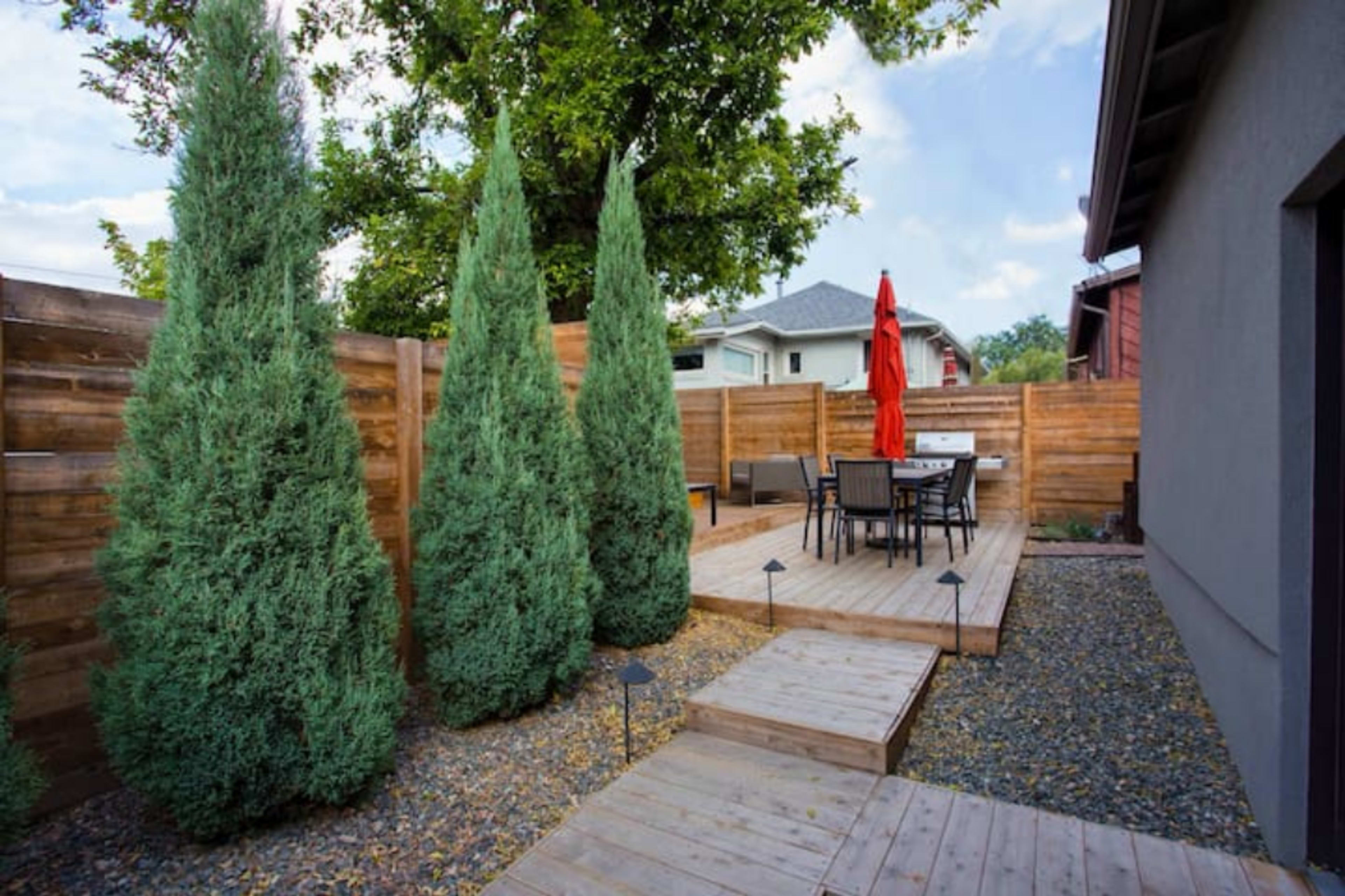 The image shows a landscaped backyard with tall, green coniferous trees, wooden fencing, a deck, and a dining set under a red umbrella.