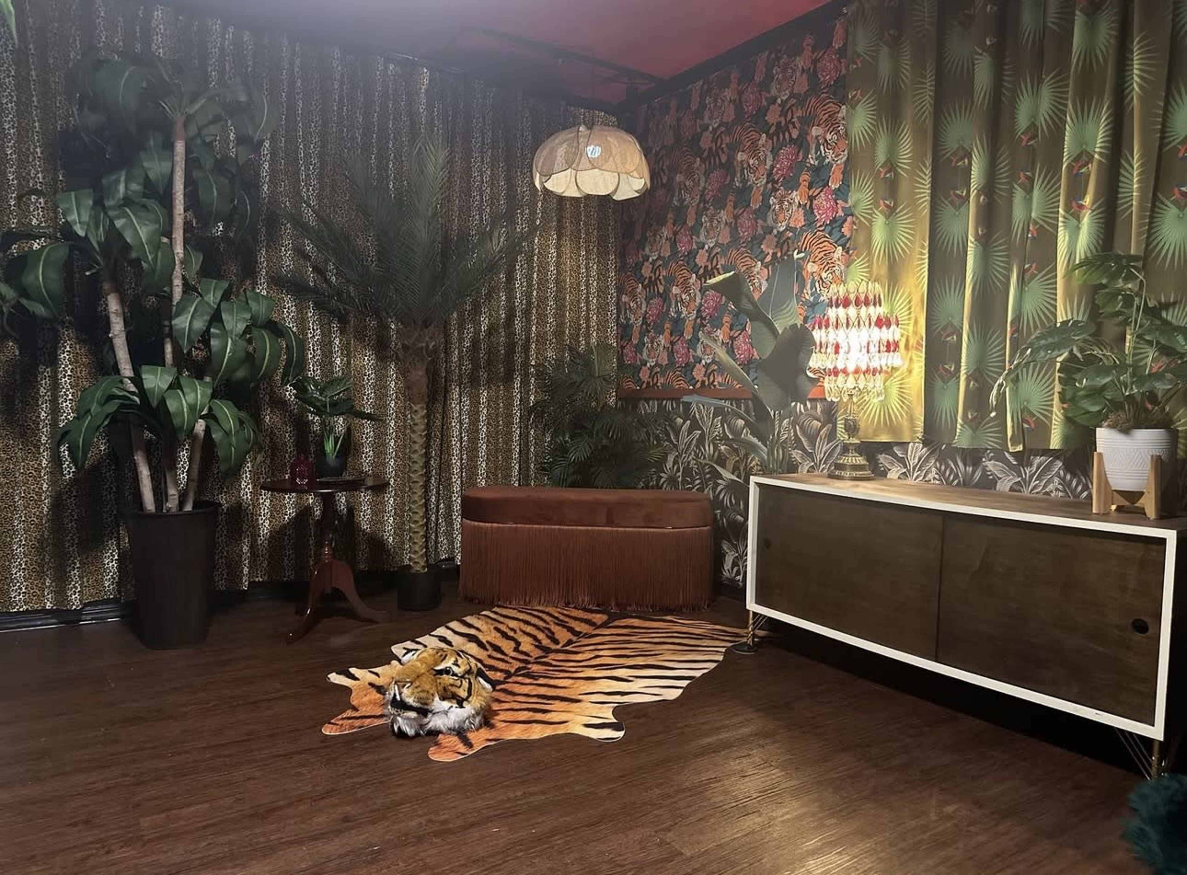 The image shows a tastefully decorated retro-themed room featuring a brown couch, a tiger-striped rug, and vibrant wallpaper with tropical patterns, surrounded by various indoor plants.