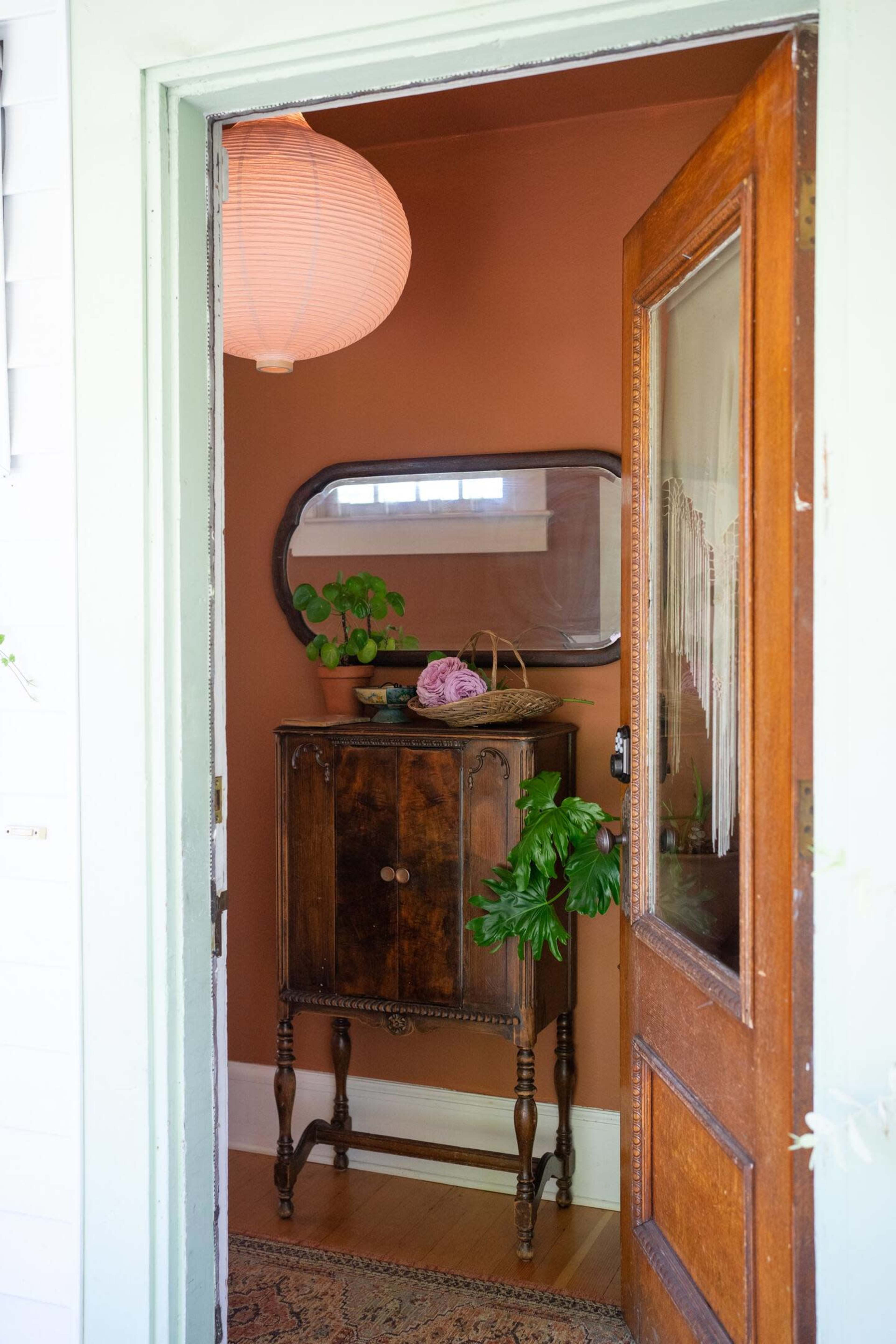 A wooden cabinet stands inside a warmly colored entryway, with a mirror above it and a pendant light hanging from the ceiling.