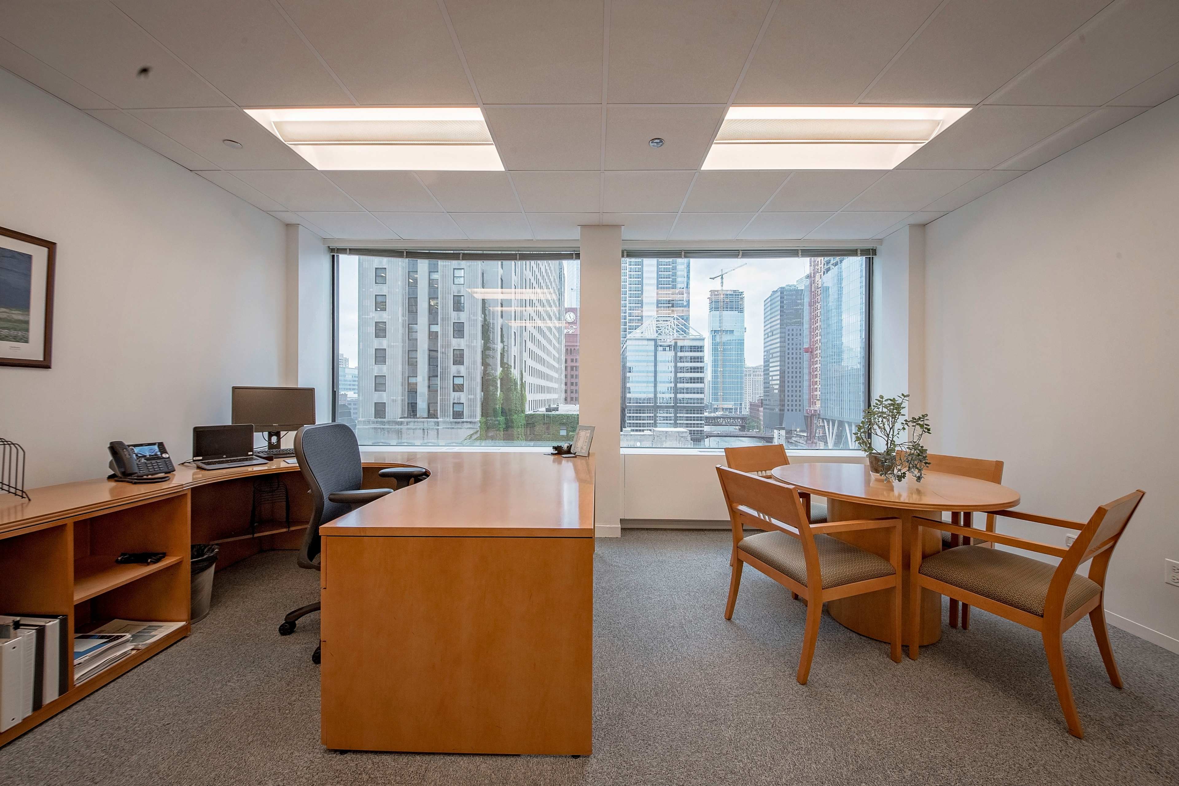 An office space features a wooden desk and chairs, with large windows displaying a cityscape.