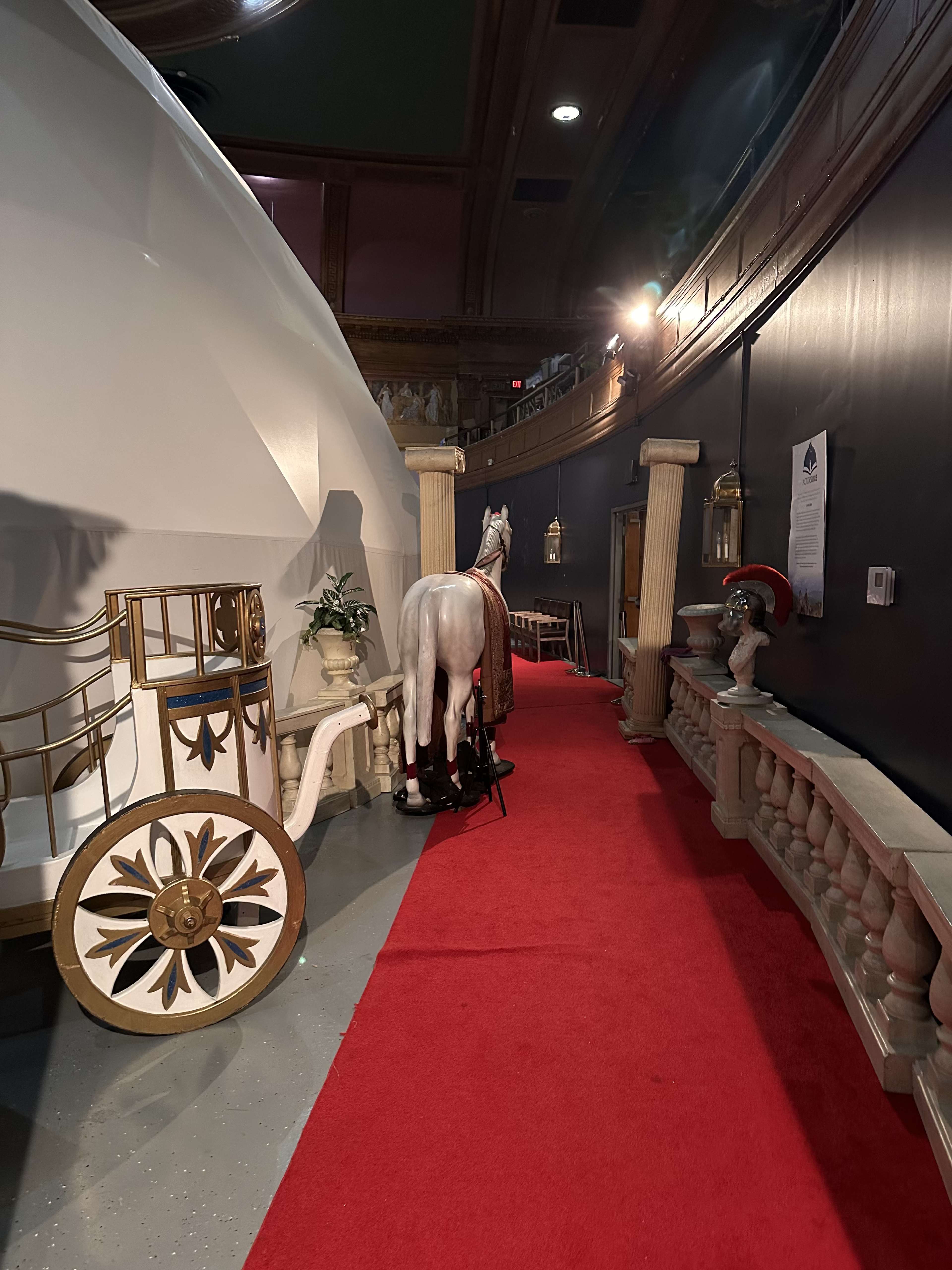 The scene features a red carpet leading alongside a wall adorned with decorative elements, including a statue and a horse figure positioned beside a decorative carriage.