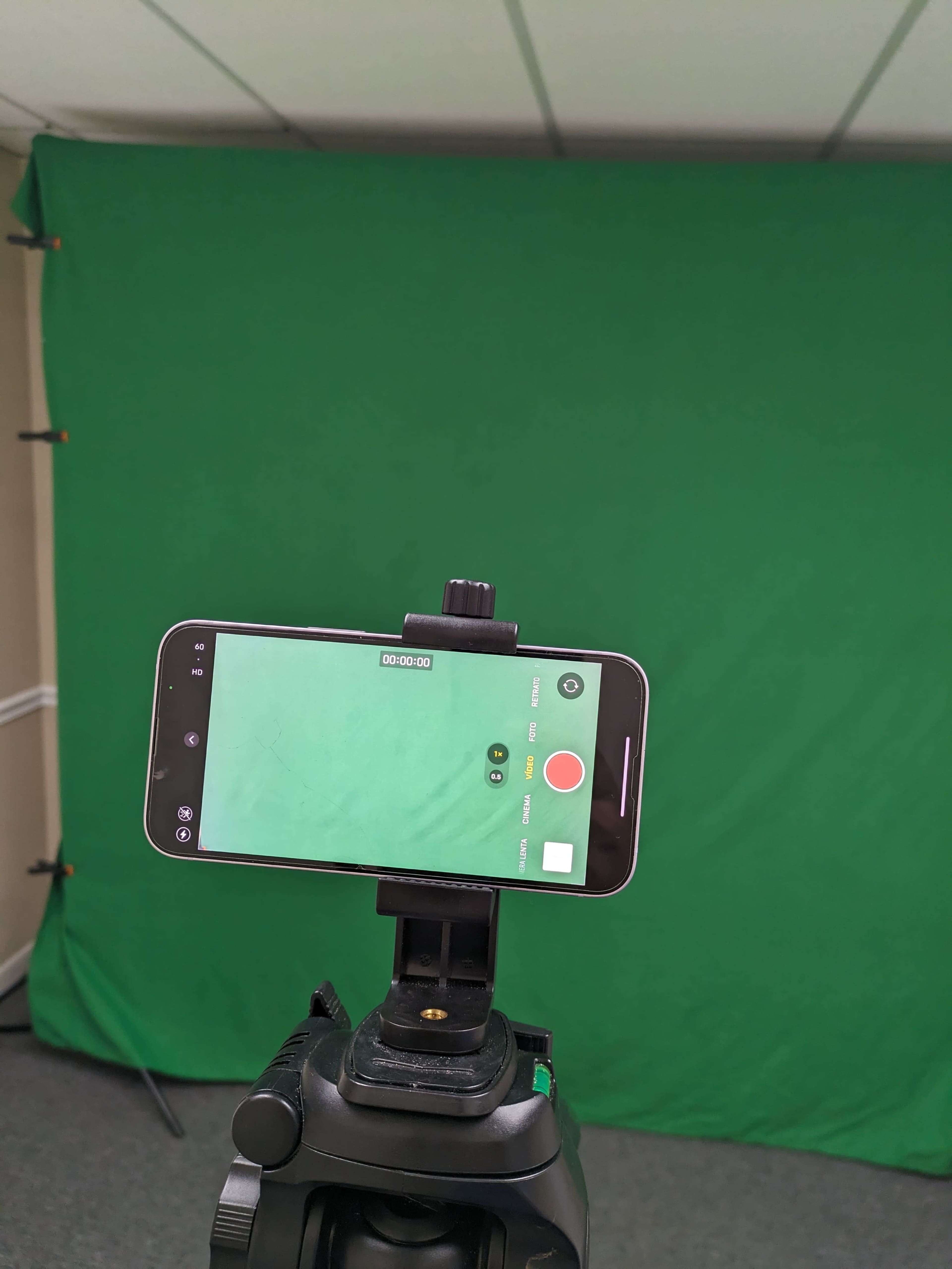 A smartphone is mounted on a tripod facing a green screen in a carpeted room.