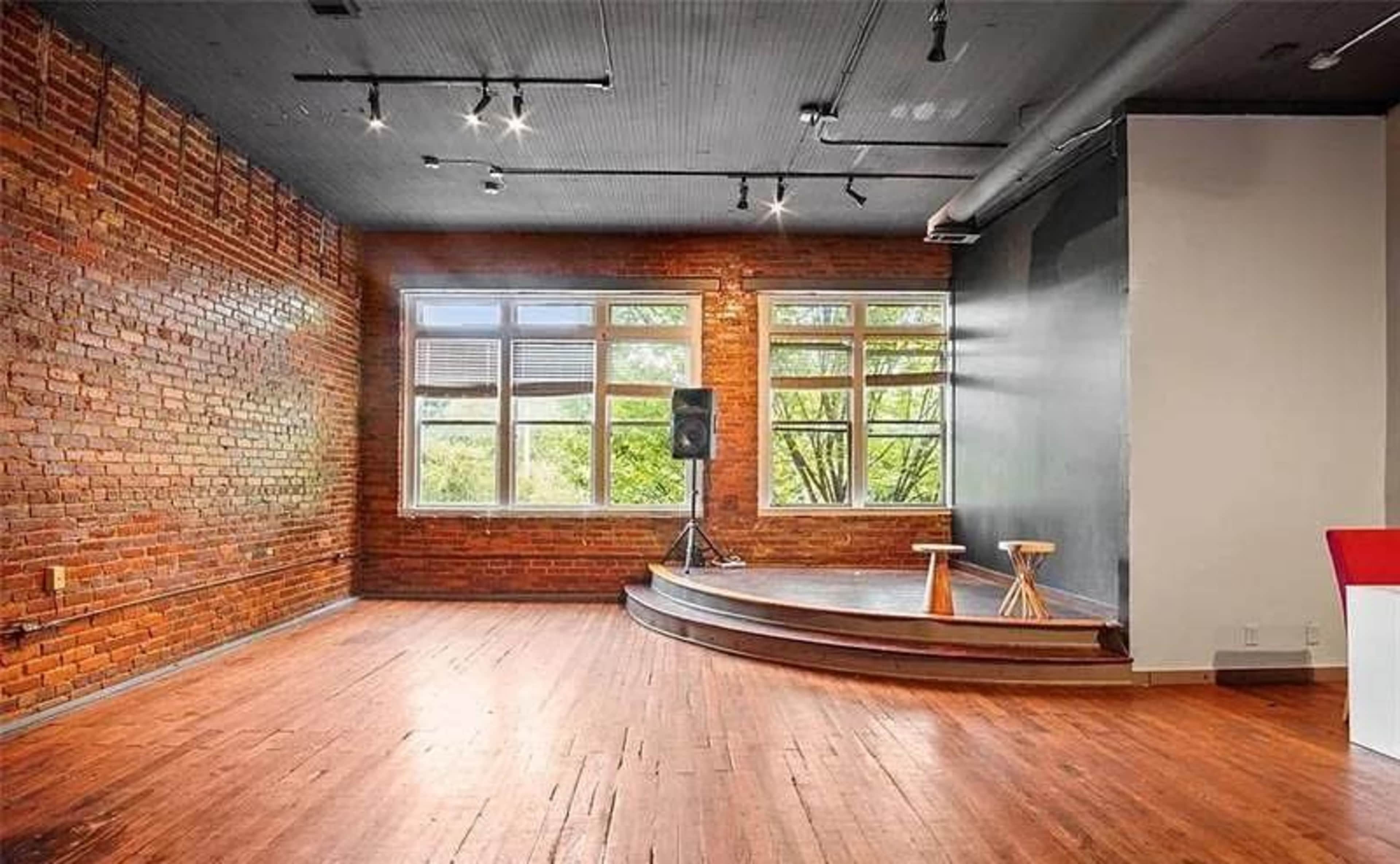 The image shows a spacious room with exposed brick walls, large windows, wooden flooring, and a small elevated stage with two stools and a speaker.
