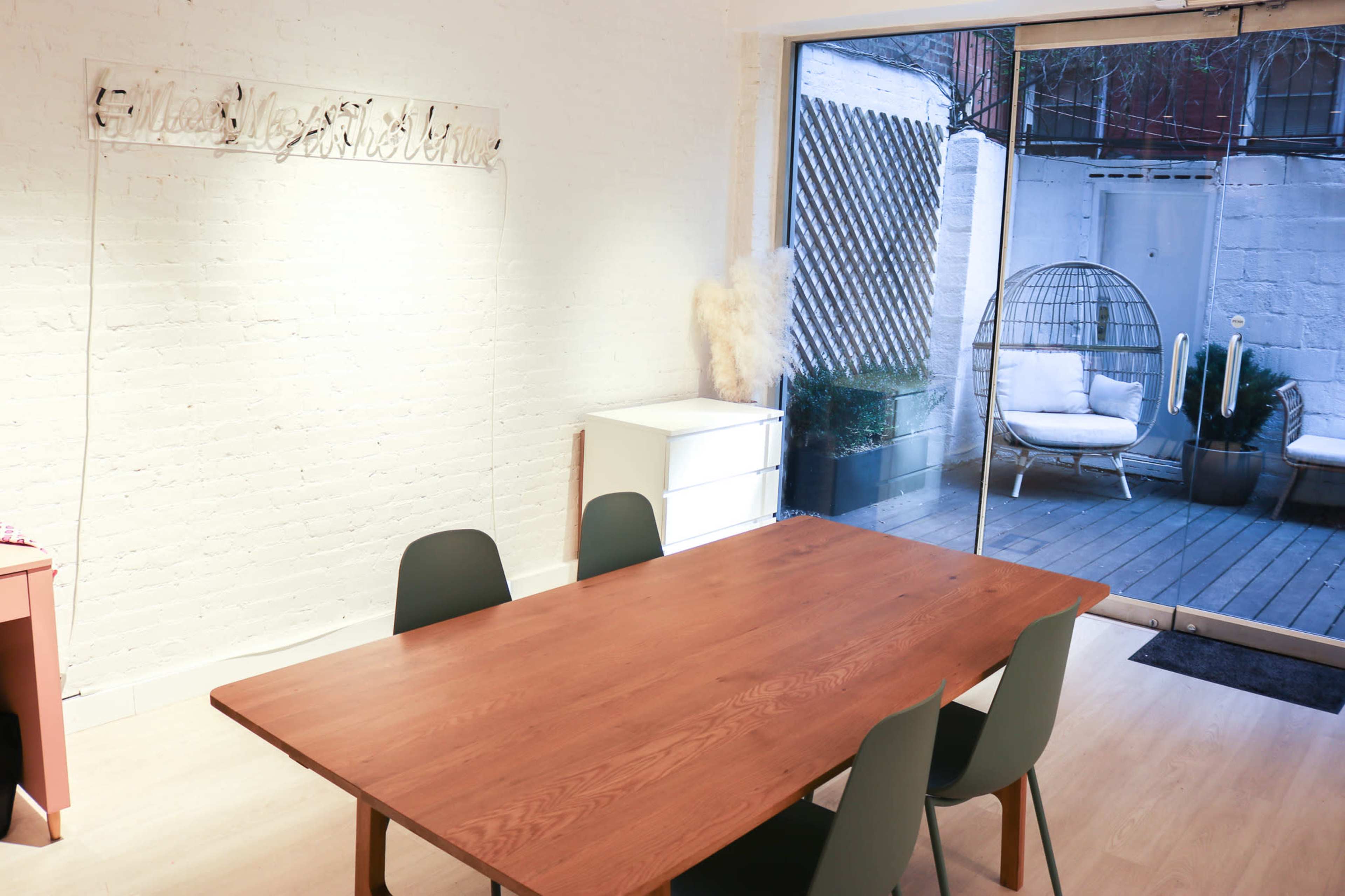 A wooden dining table surrounded by four green chairs is positioned in a bright room with a large glass door leading to a patio.