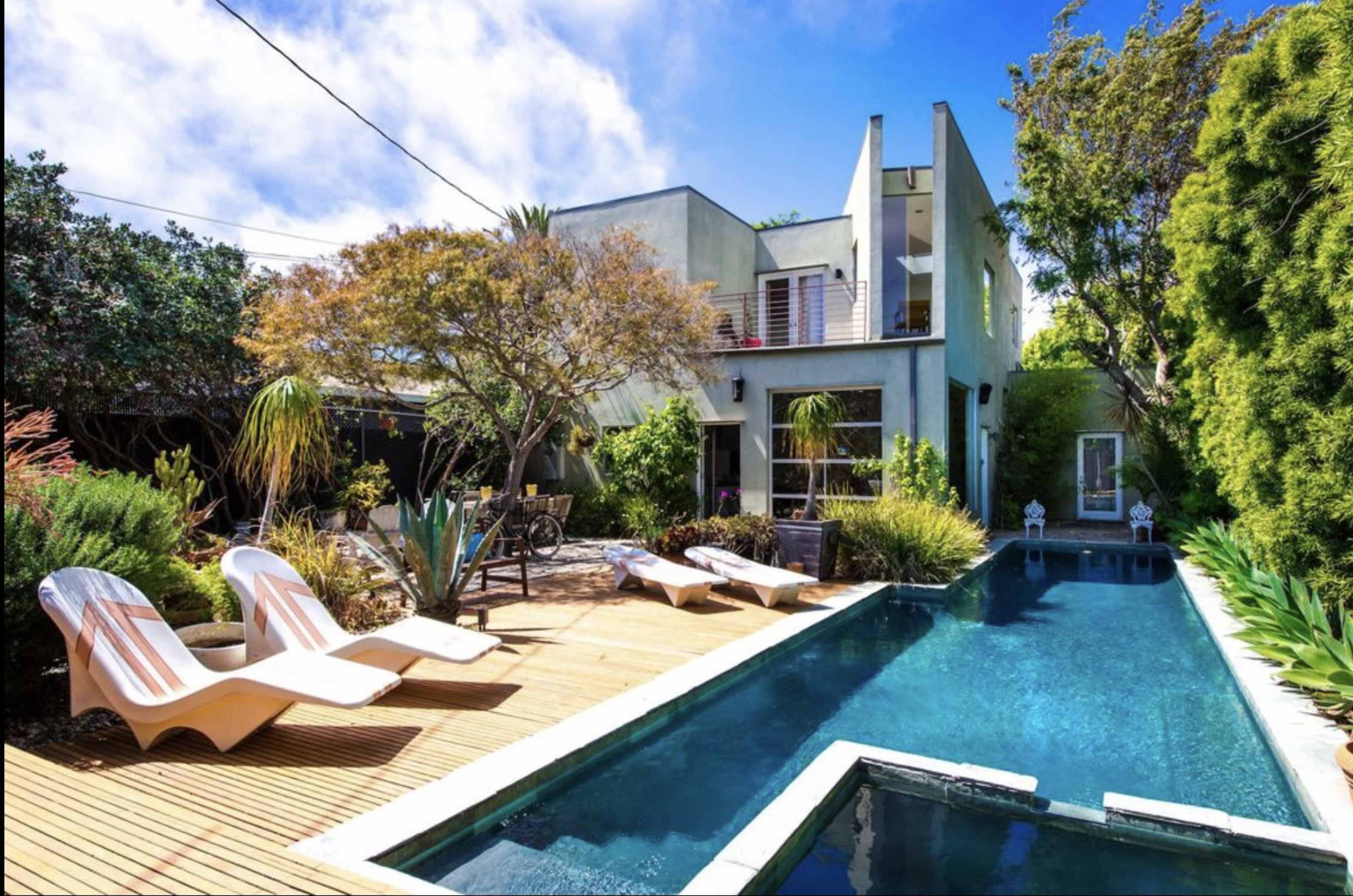 The image shows a modern house with a swimming pool and wooden decking surrounded by lush greenery.