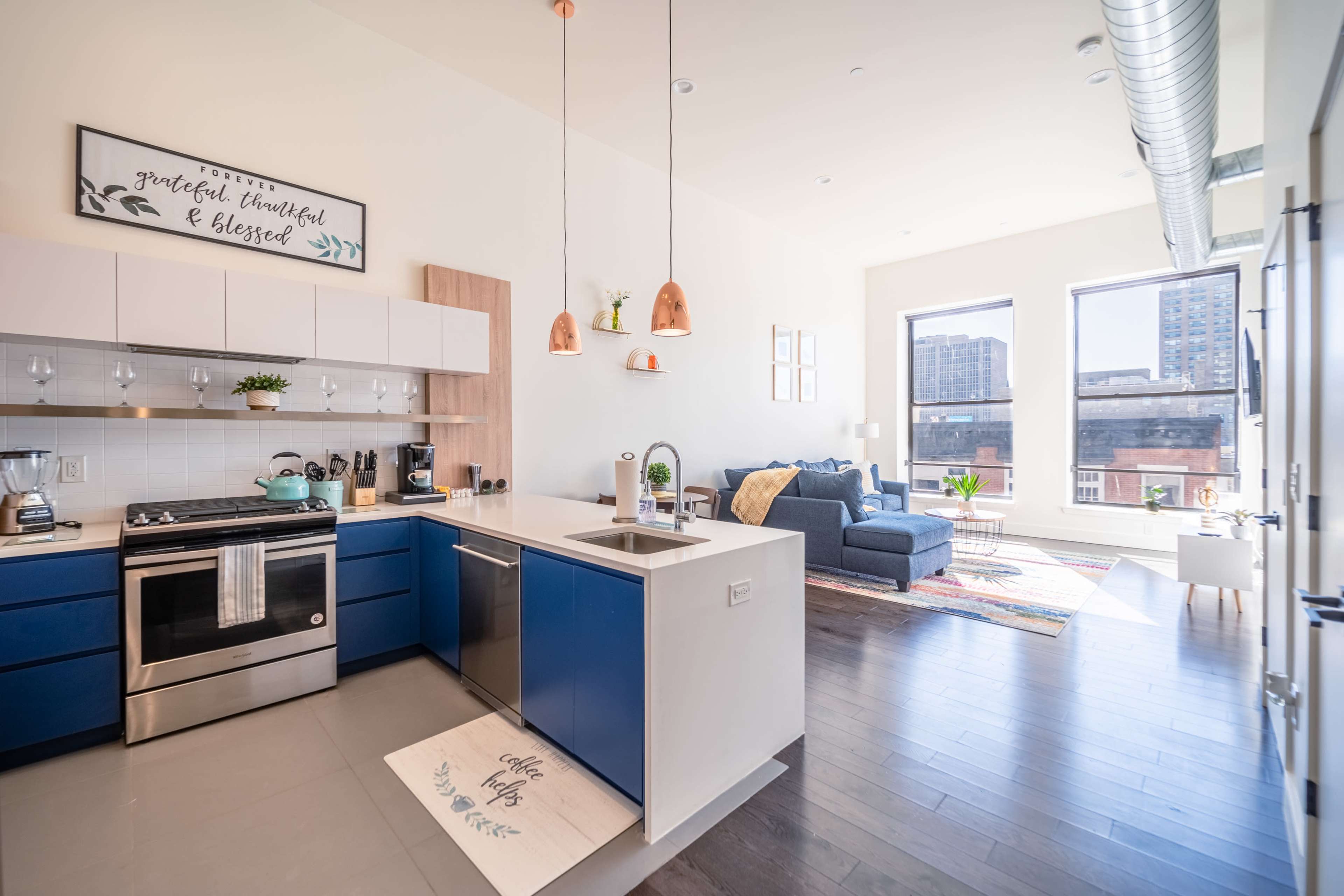 A modern kitchen with blue cabinets, copper pendant lights, and an open living area featuring large windows and a cozy seating arrangement.
