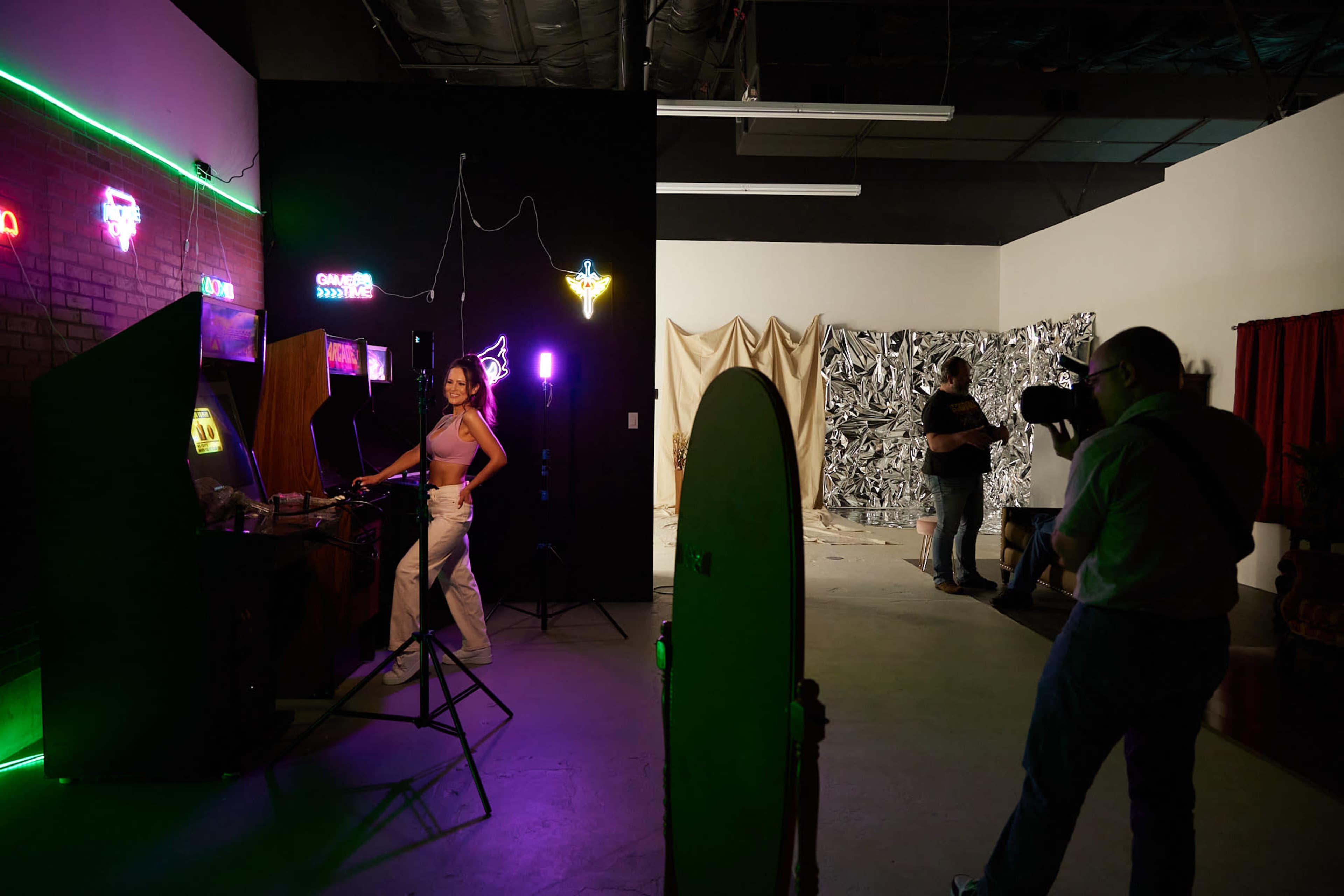 A woman poses near a retro arcade game in a studio filled with colorful lights and photography equipment while another person captures the scene with a camera.